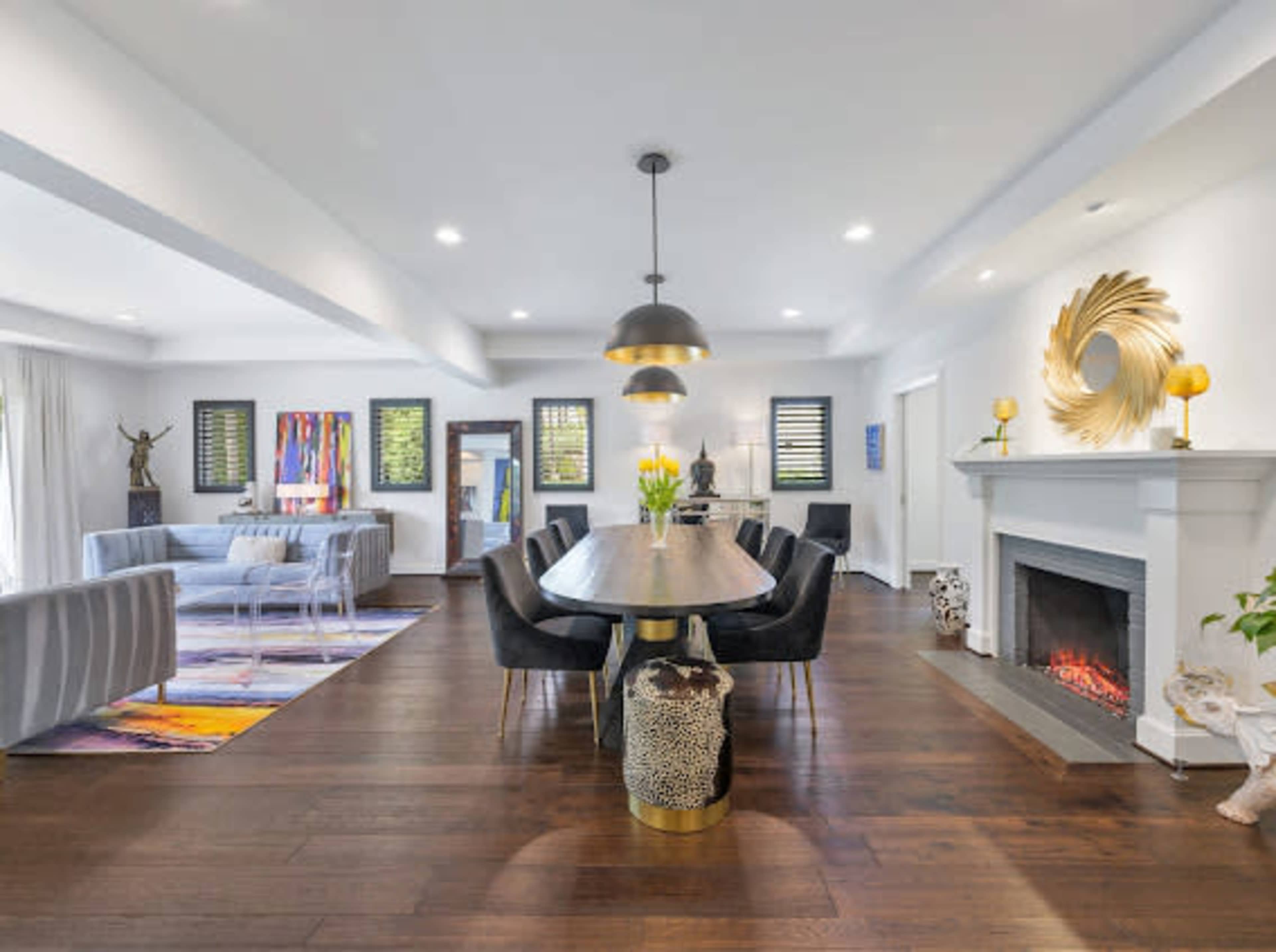 A dining area features a long table surrounded by black chairs, with a fireplace and modern decor in a spacious, well-lit room.