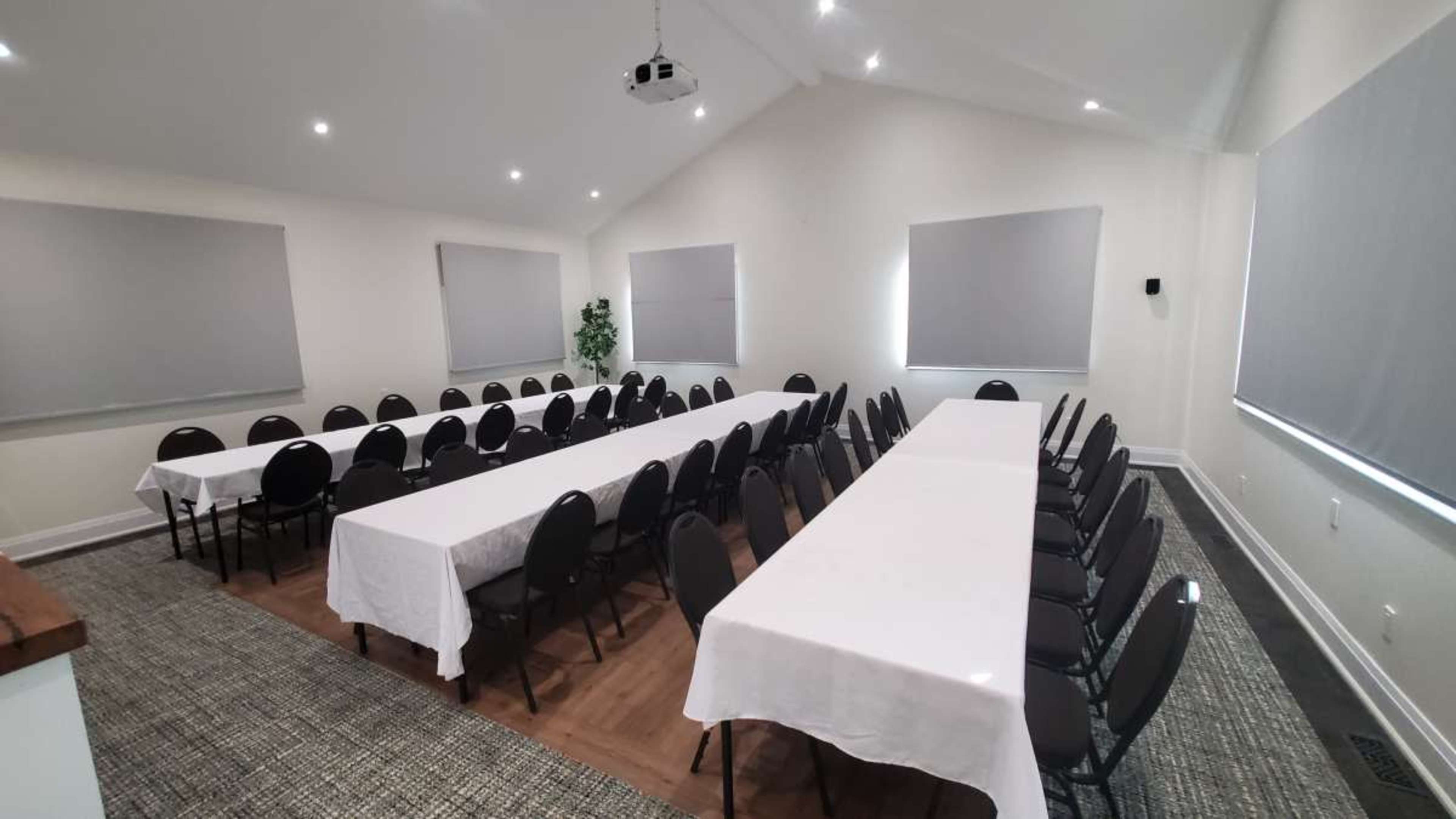 A spacious room is set up with multiple long tables, covered in white tablecloths, arranged for a meeting or event.