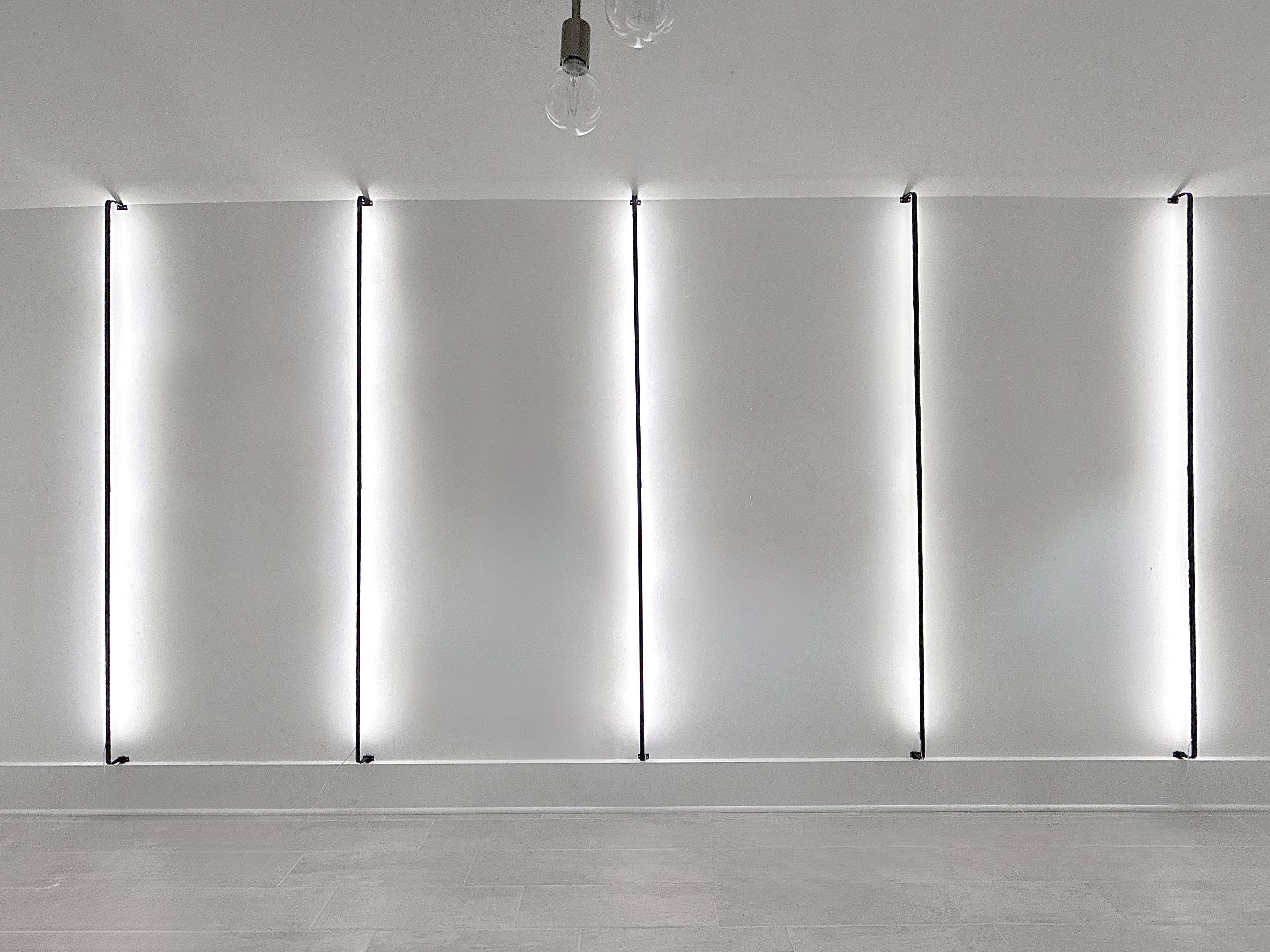The image shows a plain white wall with four vertical strips of light illuminating the surface.