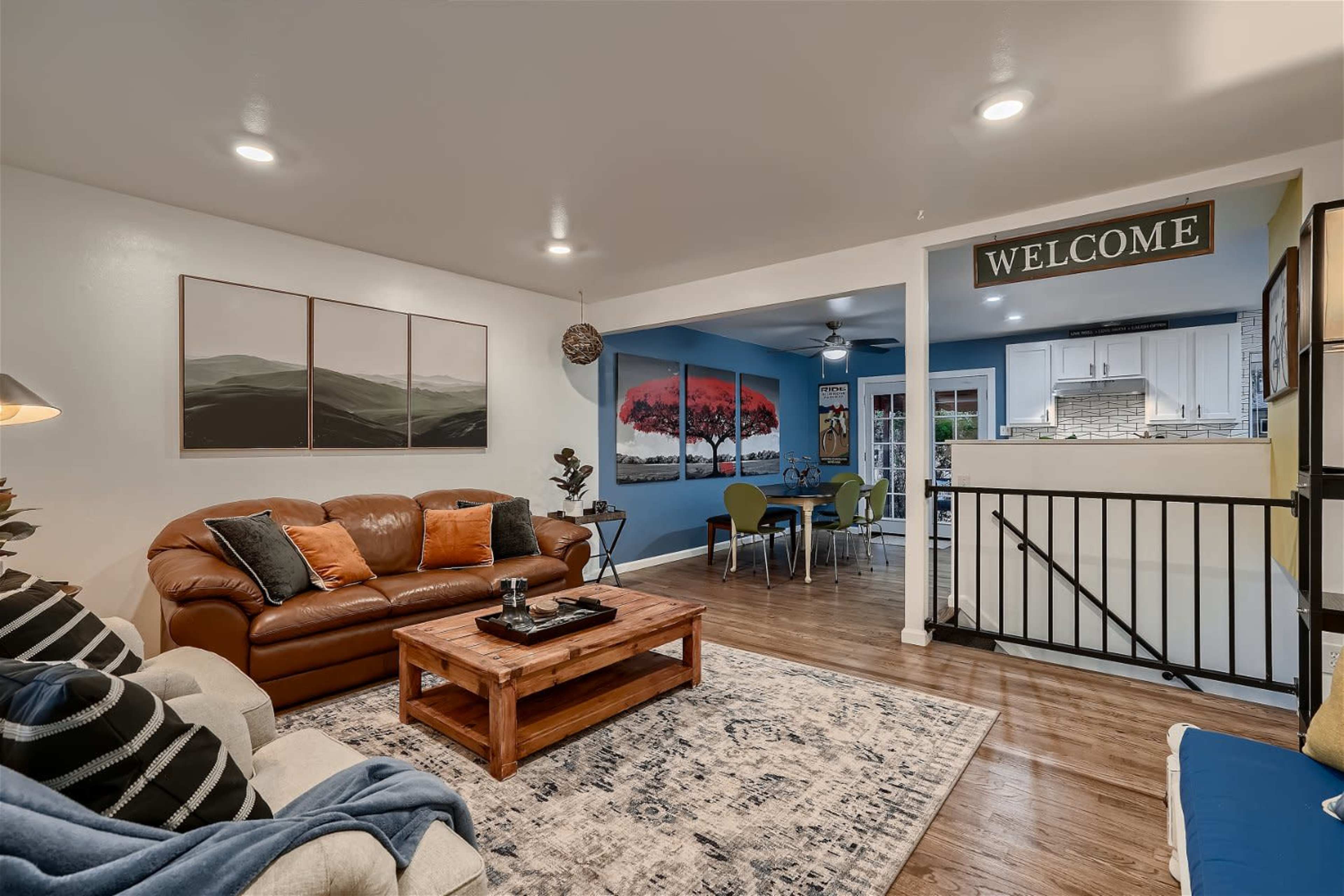 The image shows a cozy living room with a brown leather sofa, wooden coffee table, and a welcoming sign, leading to a dining area decorated with blue walls and artwork.