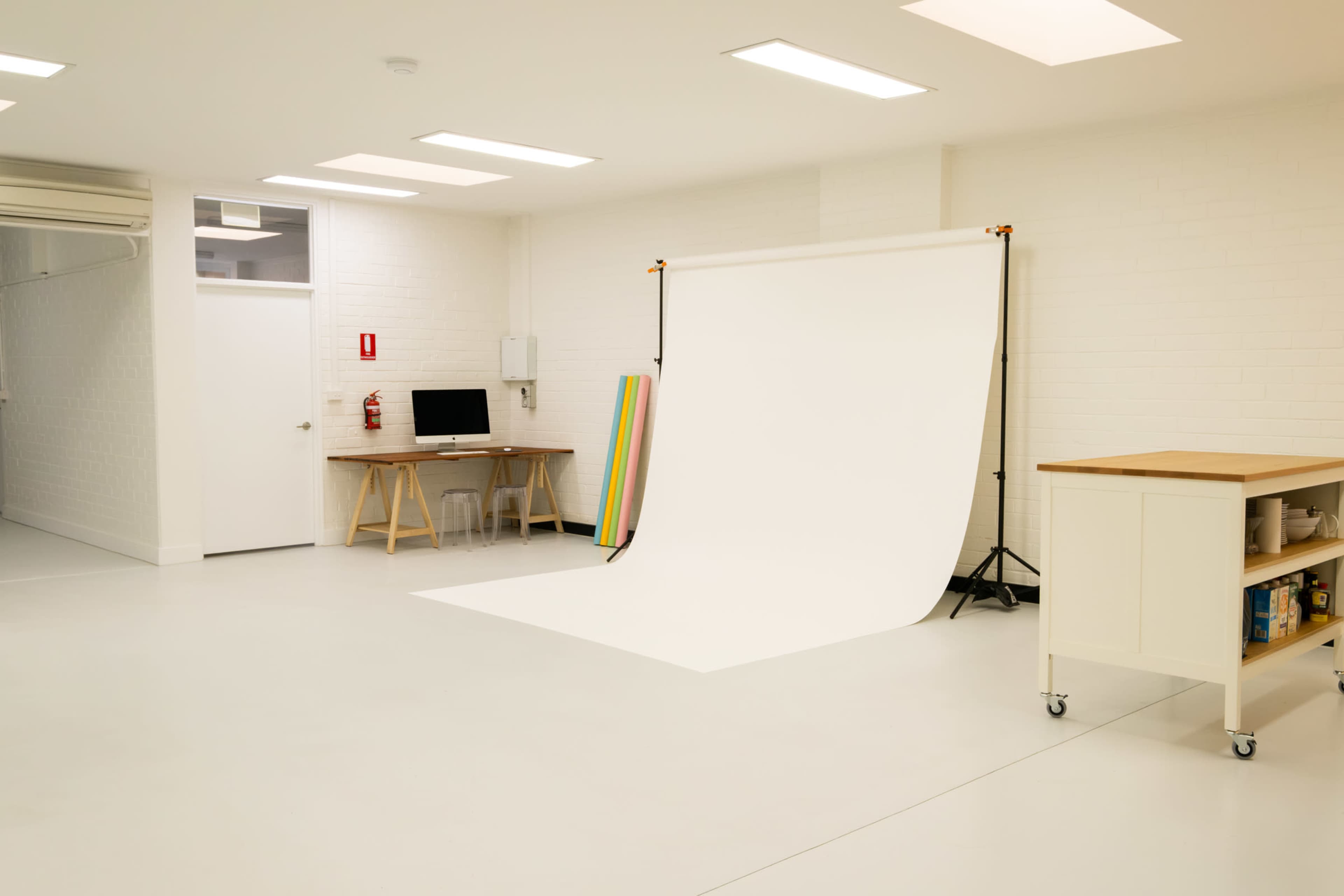 The image shows a clean and well-lit studio space featuring a white backdrop, a desk with a computer, and a rolling cart with supplies.