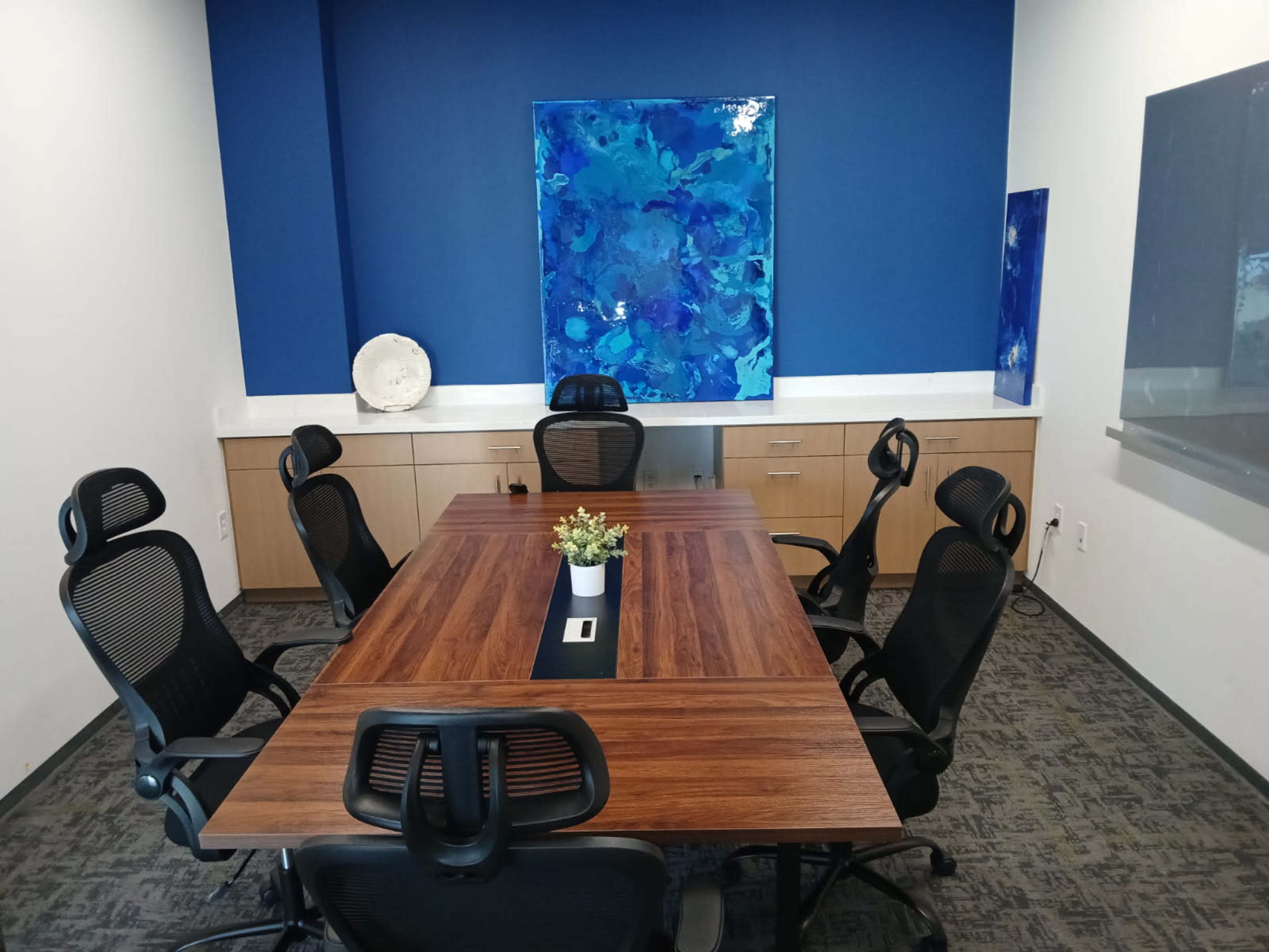 A conference room features a large wooden table surrounded by six ergonomic chairs, with blue artwork hanging on the wall and cabinetry in the background.