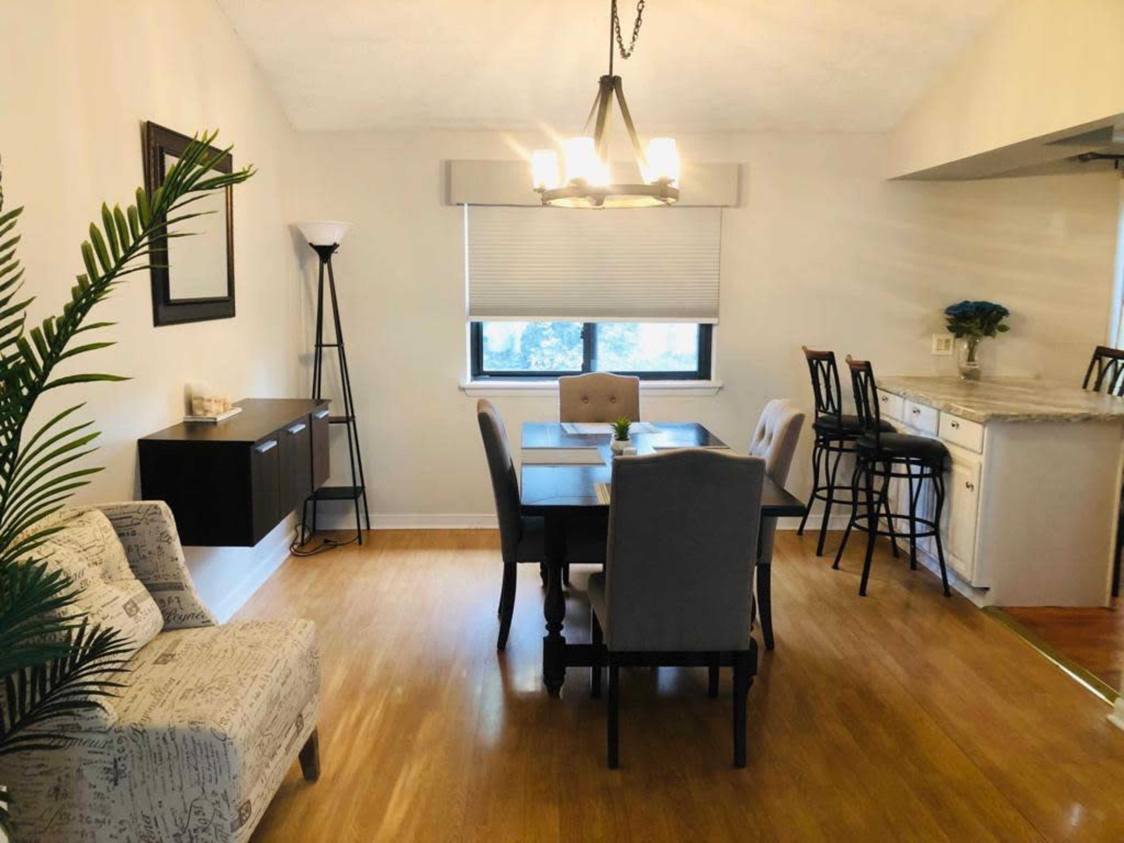 A dining area with a table, chairs, and a modern light fixture, adjacent to a kitchenette.