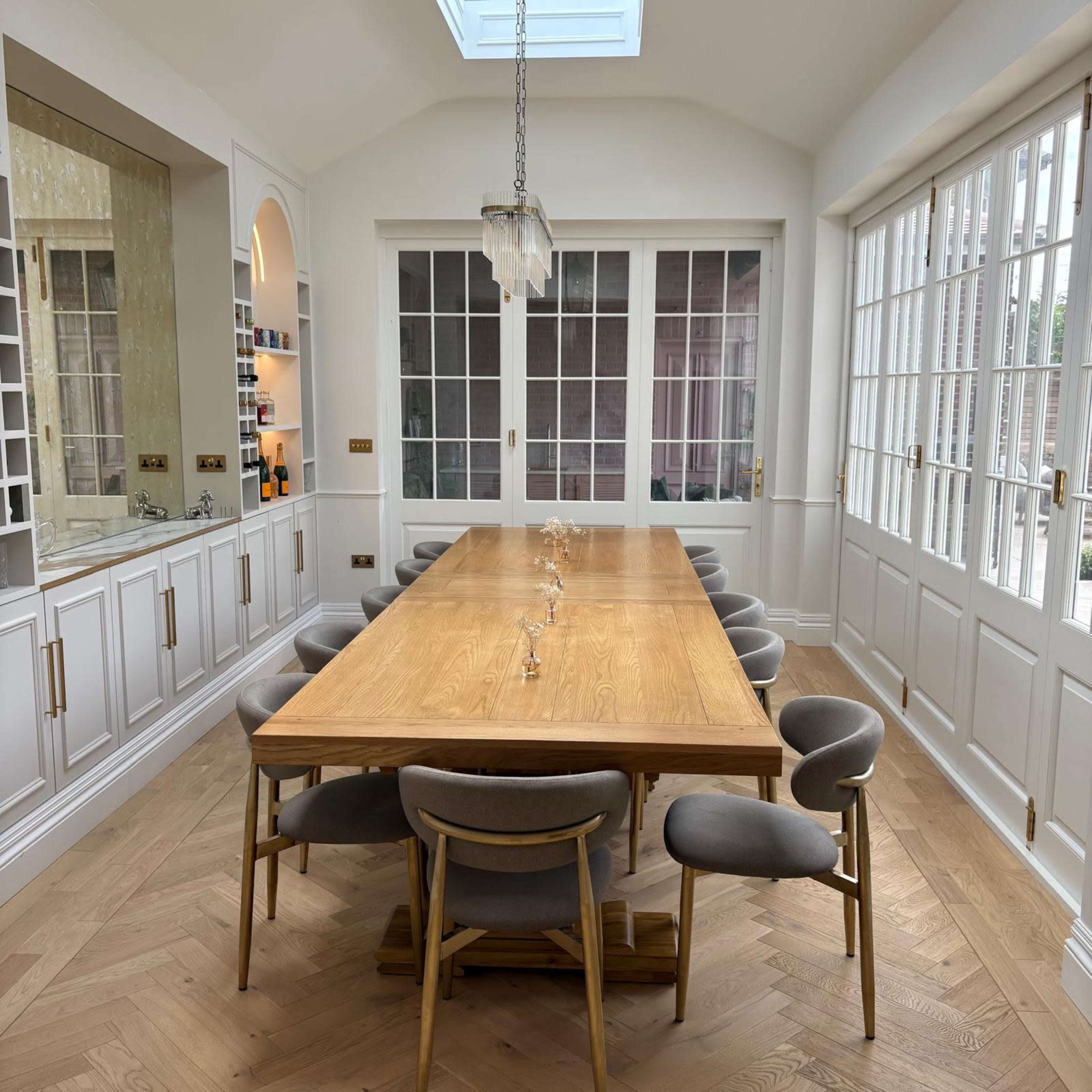 The image shows a spacious dining area with a long wooden table surrounded by upholstered chairs, set against a backdrop of large glass doors that let in natural light.
