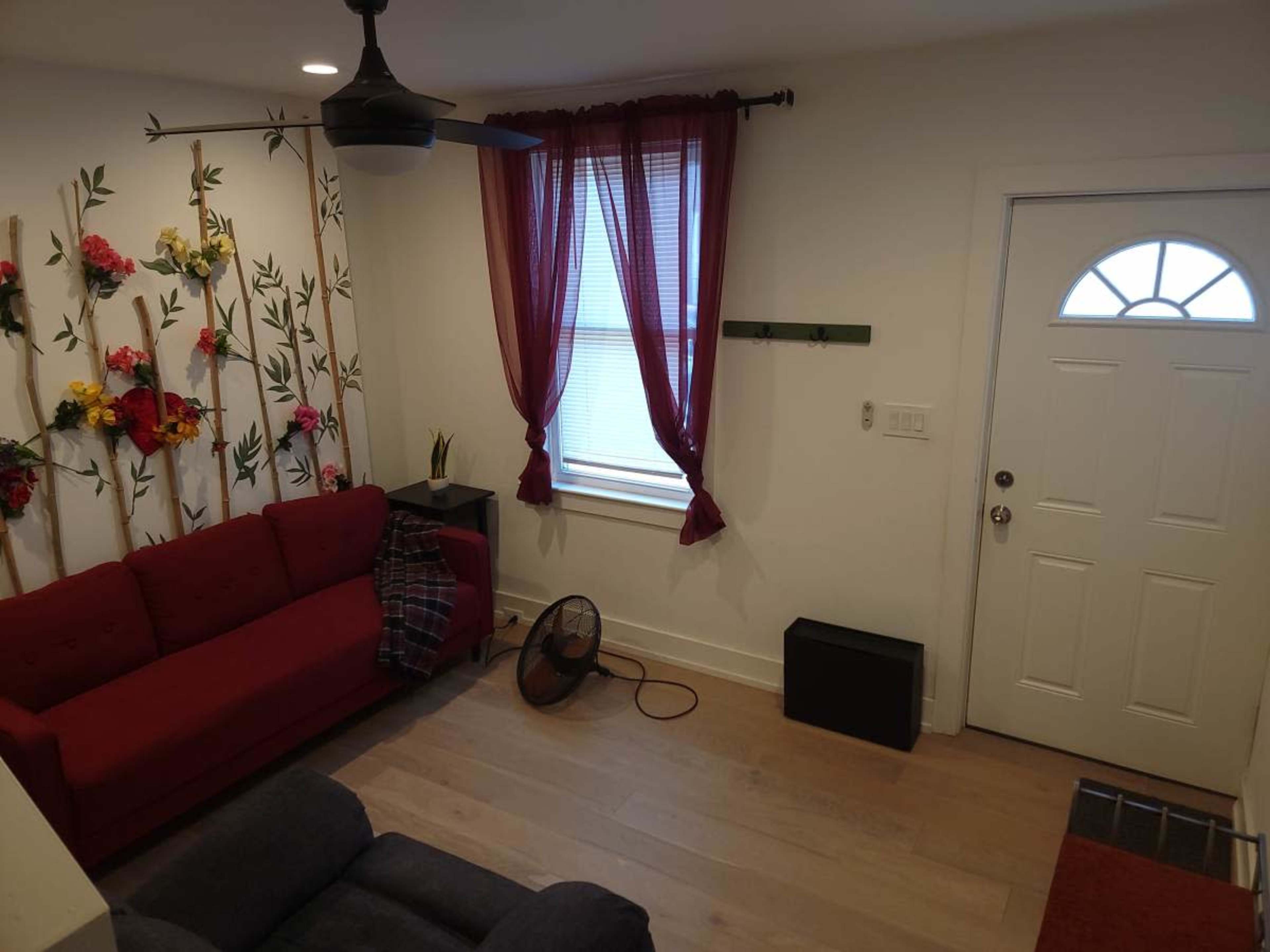 The image shows a small living room with a red couch, a decorative wall with flowers, a fan on the floor, and a door leading outside.
