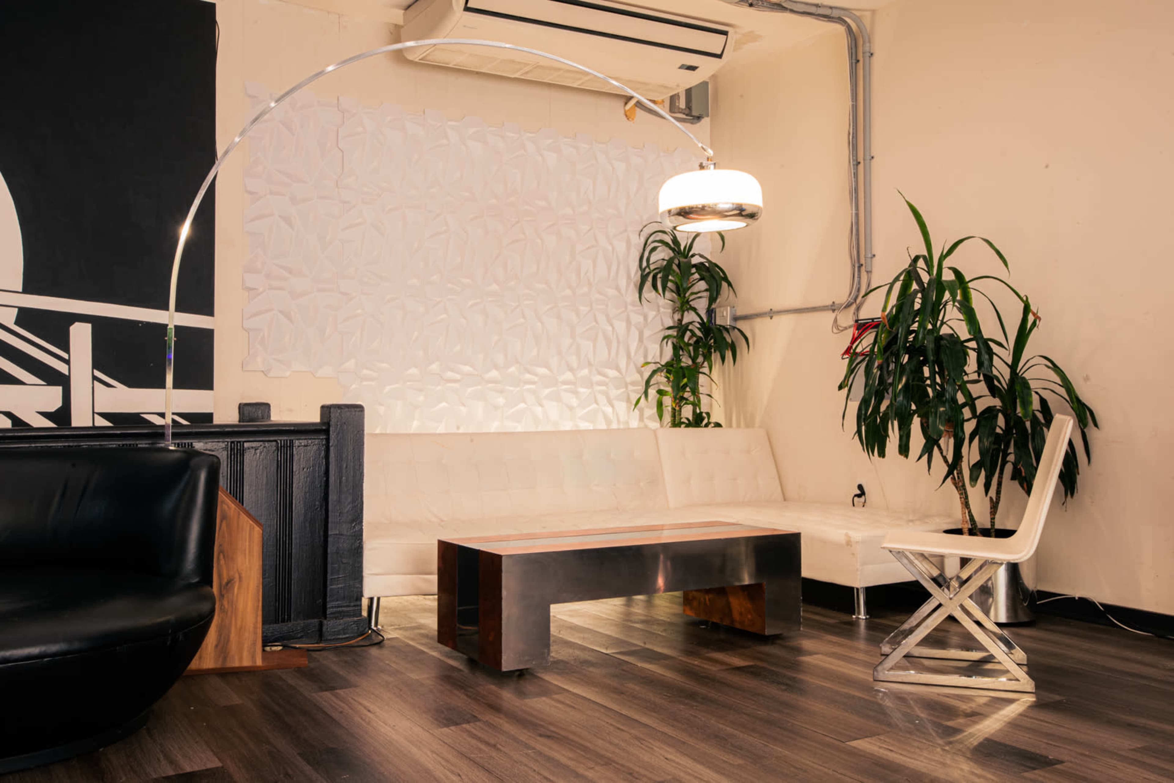 The scene features a modern lounge area with a white leather sofa, a metal coffee table, a floor lamp, and indoor plants against a textured wall.