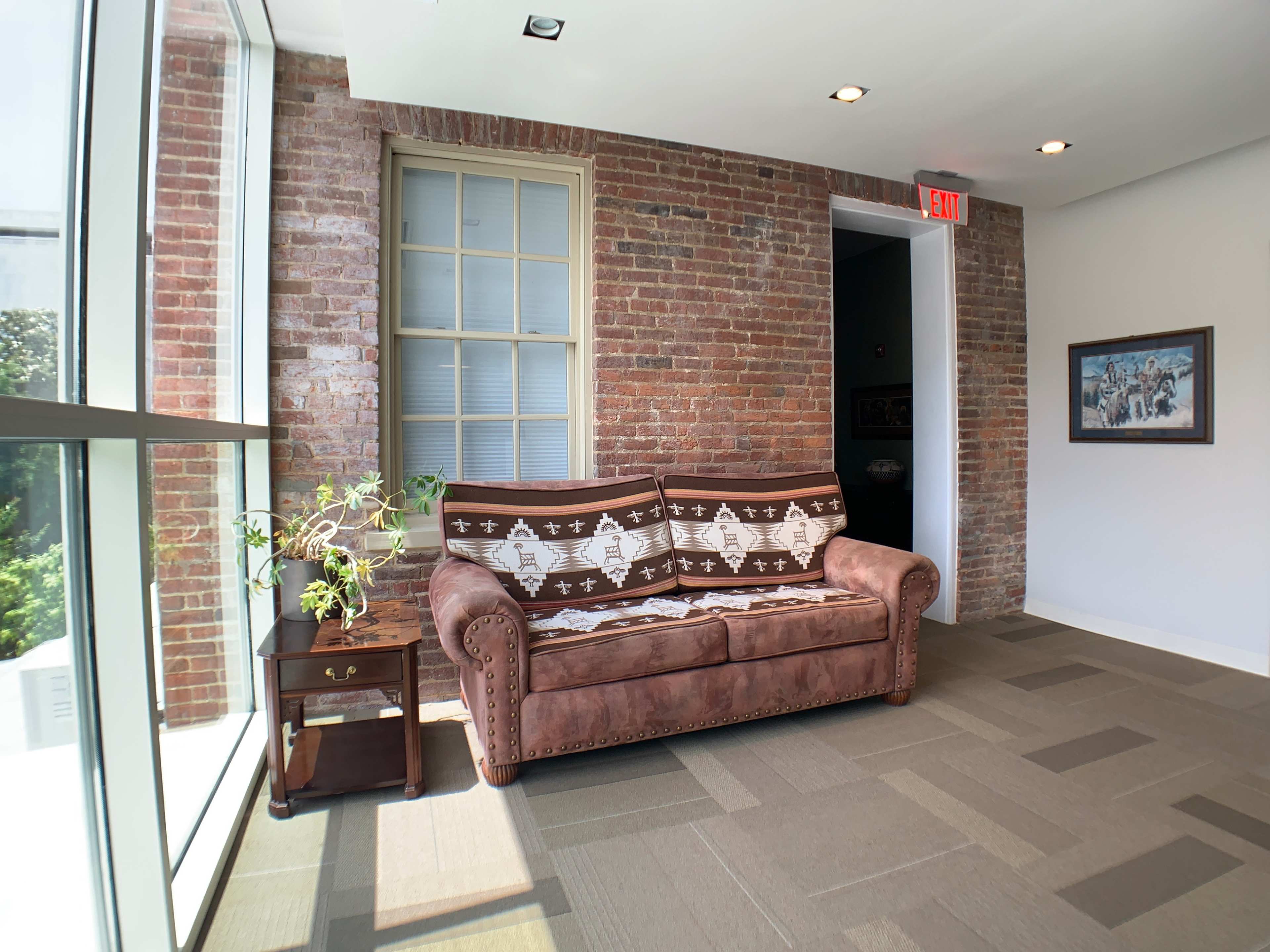 A brown sofa with a patterned throw placed against a brick wall next to a small table and a large window.