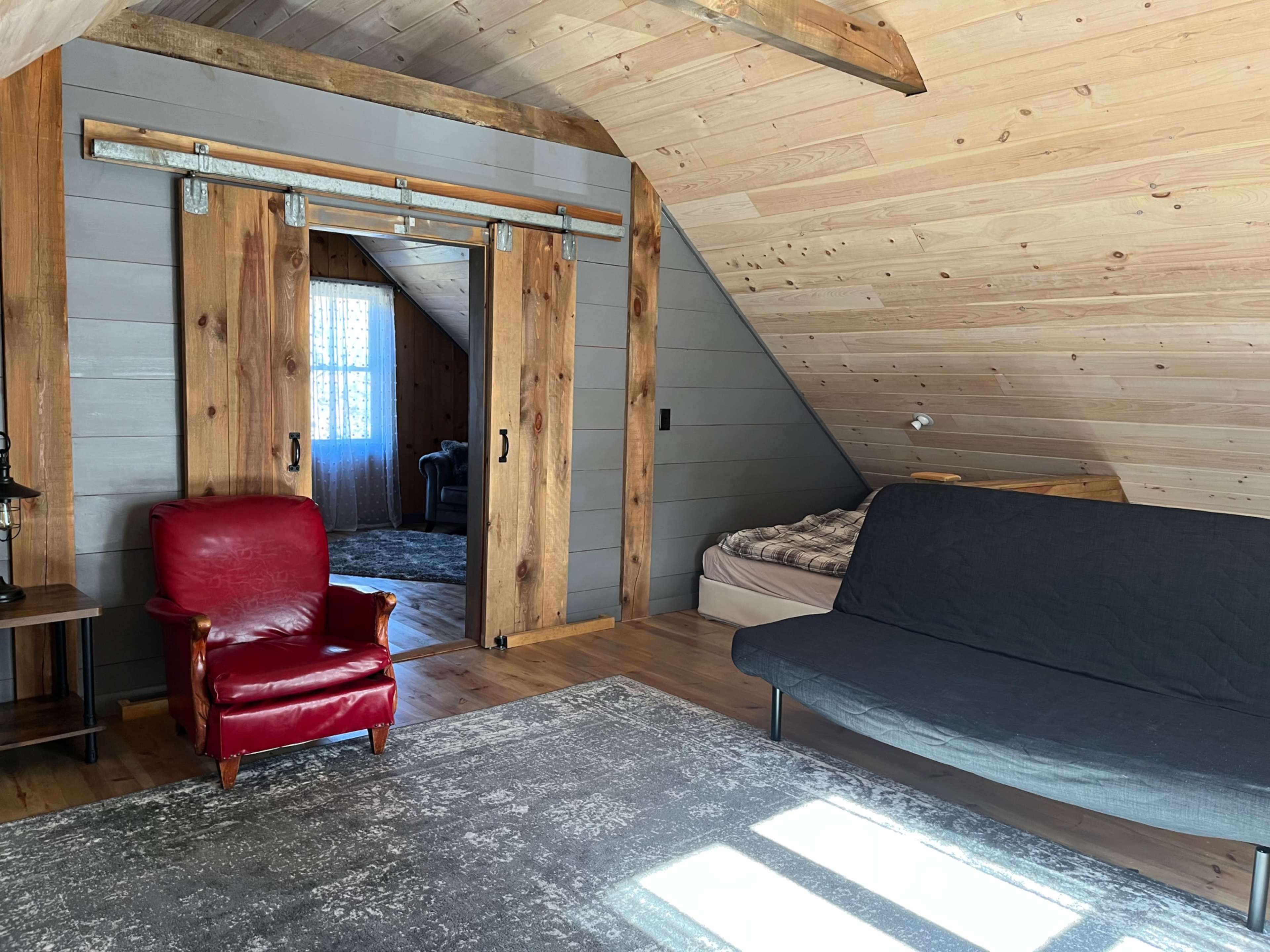 An attic room featuring a red armchair, a dark sofa, a bed against the wall, and wooden accents in the ceiling and walls.