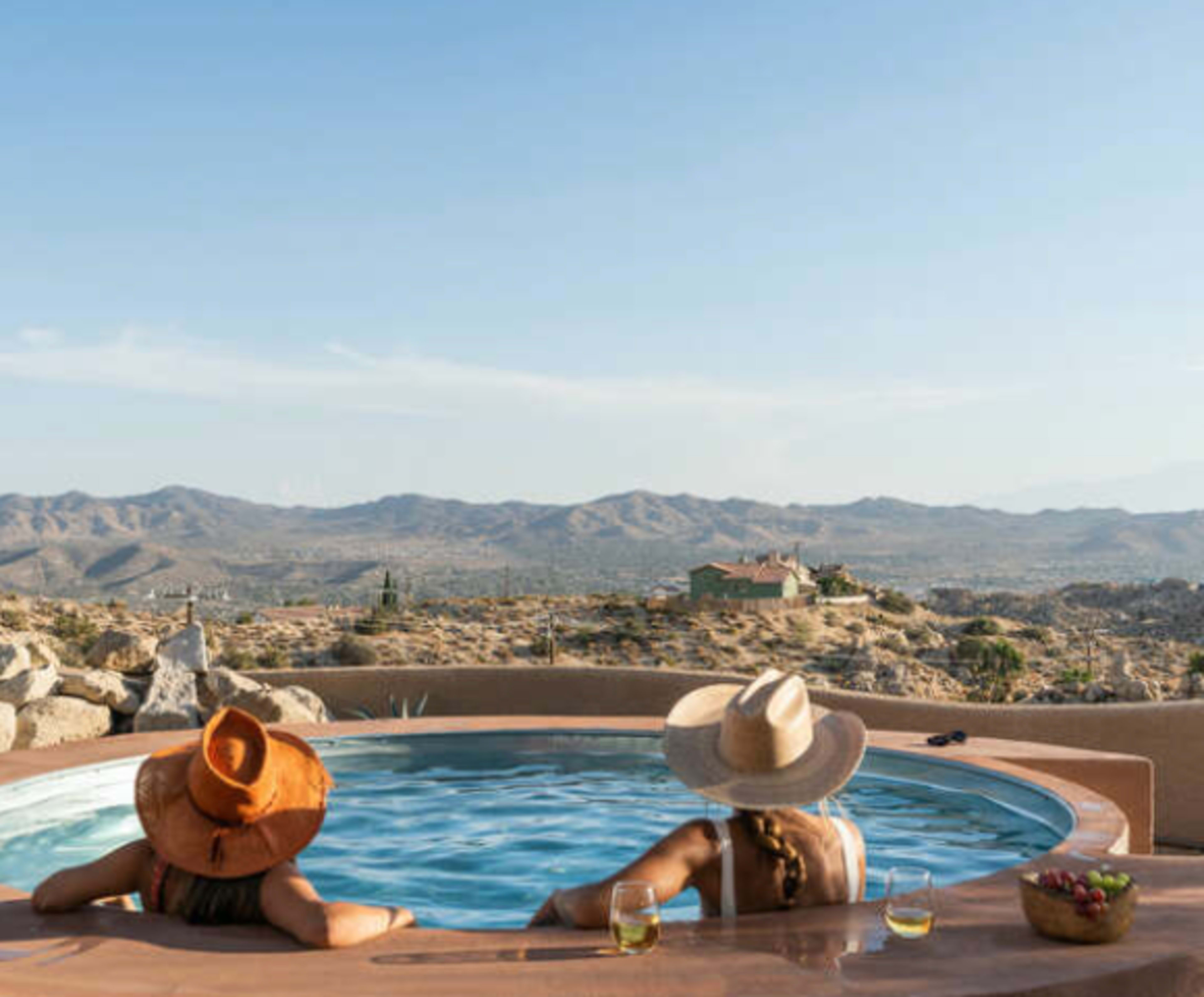 Two people wearing hats relax in a hot tub, overlooking a mountainous desert landscape with a clear sky.