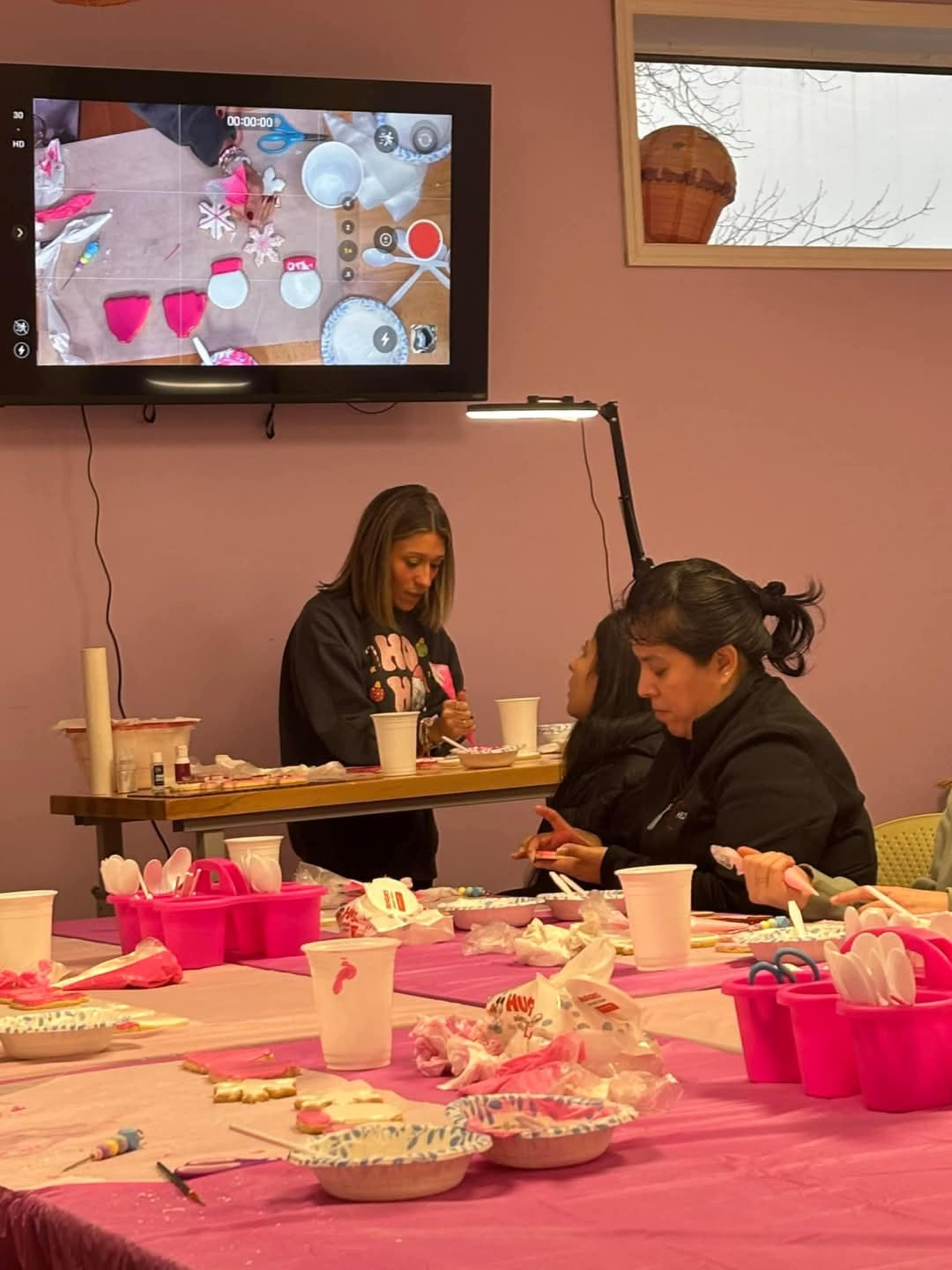 The image shows a group of people participating in a crafting activity in a colorful, well-lit room with tables covered in craft supplies.