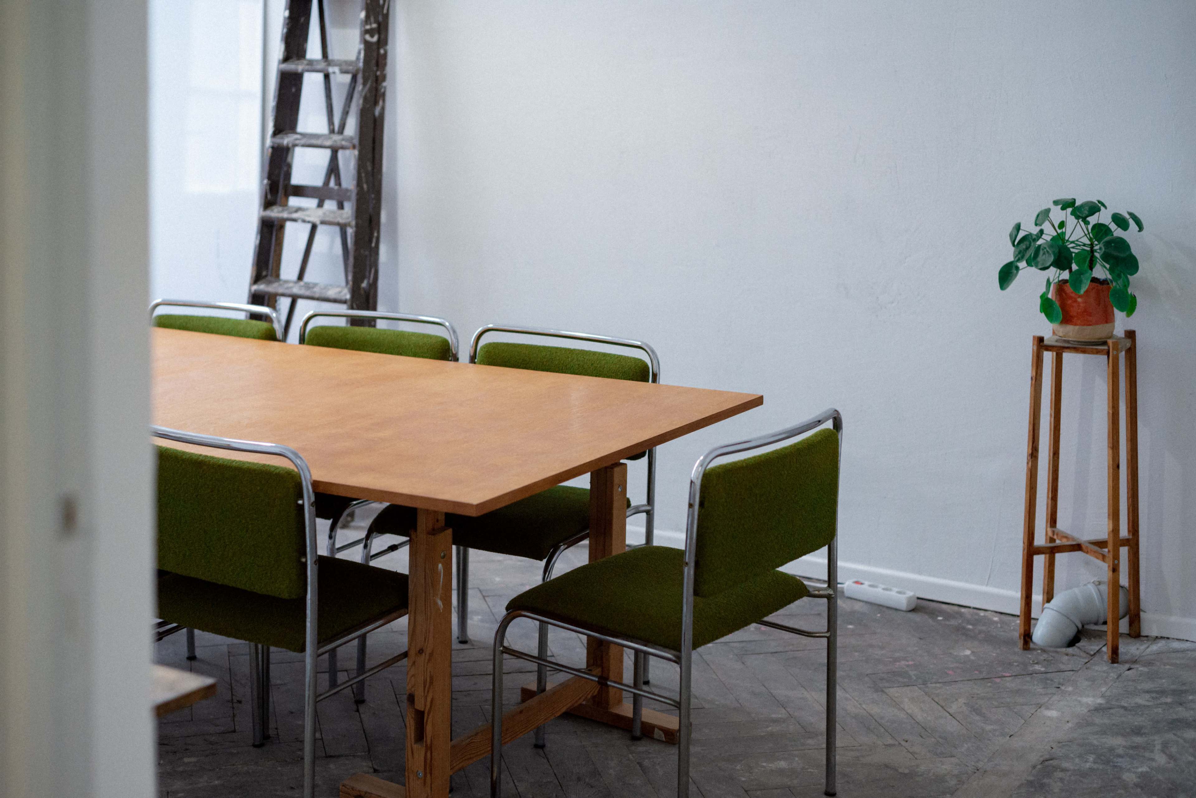 A wooden table with green chairs is set in a sparsely furnished room, featuring a ladder against the wall and a potted plant on a stand.
