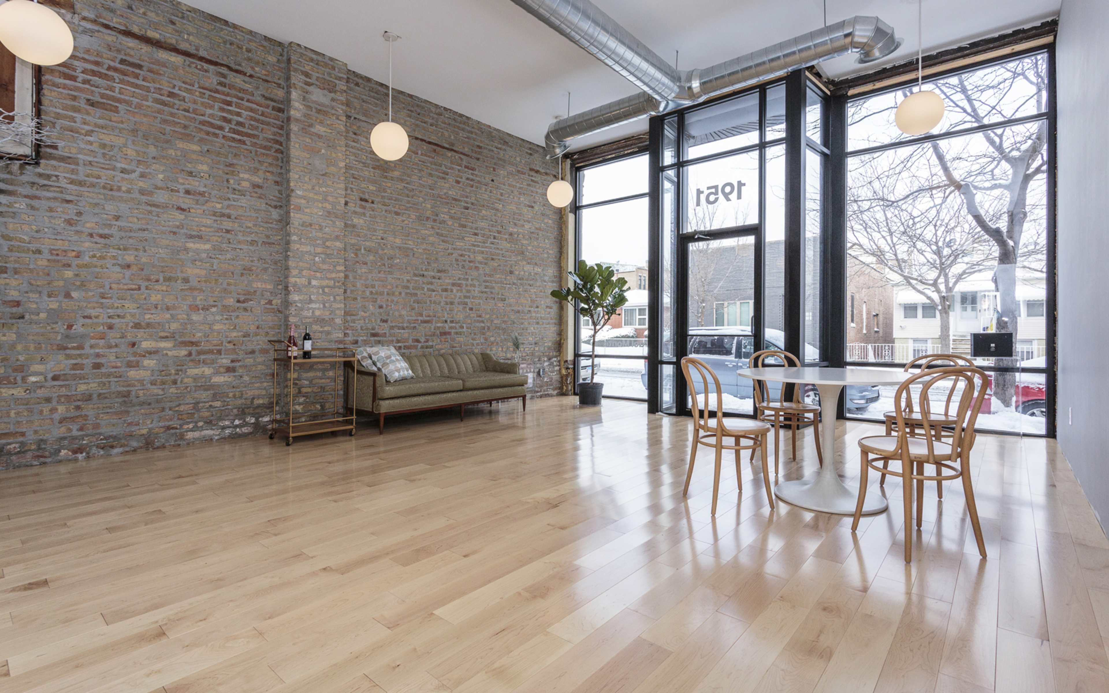 A modern, open interior space with hardwood floors, a brick wall, a sofa, a small dining table with chairs, and large windows facing the street.