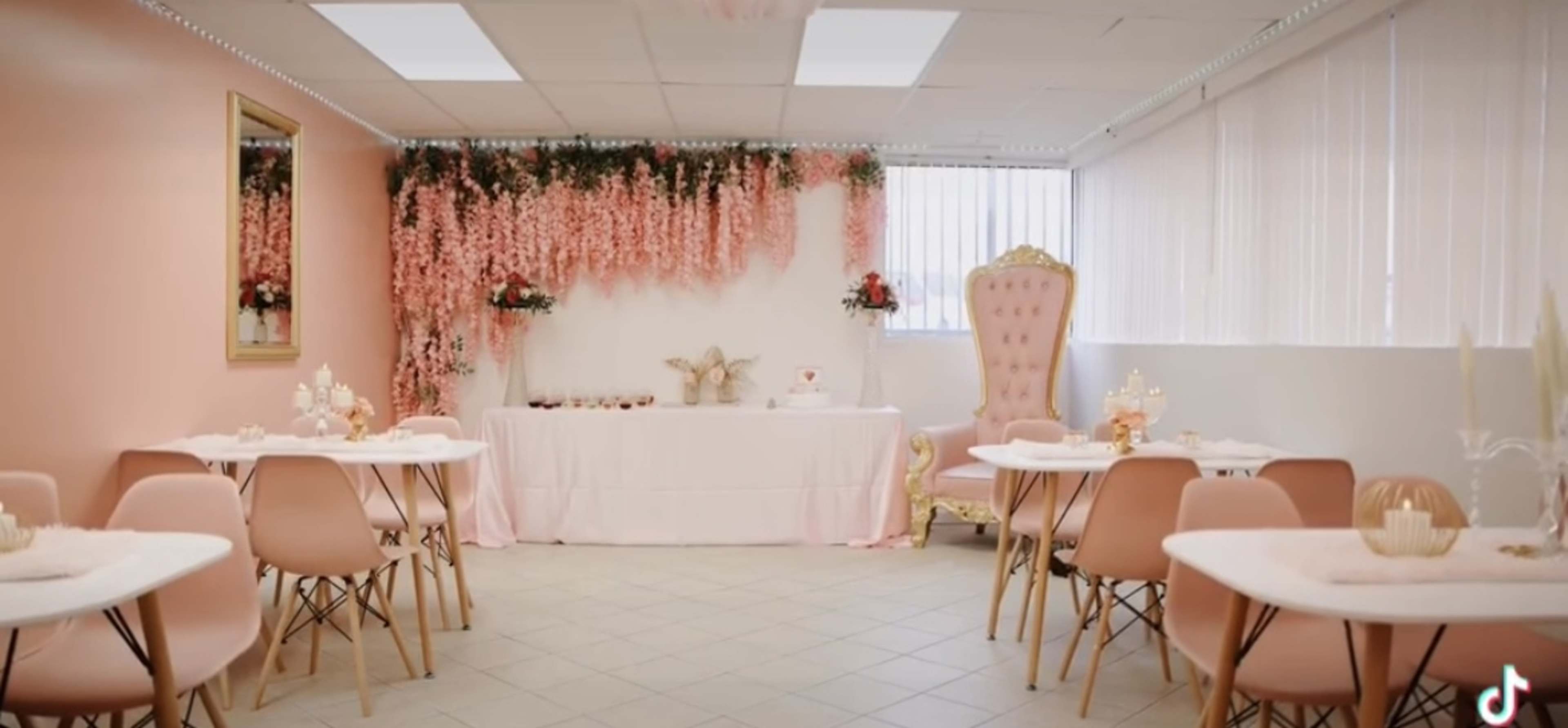The image shows a decorated indoor space with pink walls, floral drapery, white tables, and elegant chairs arranged around a central table adorned with decorative items.