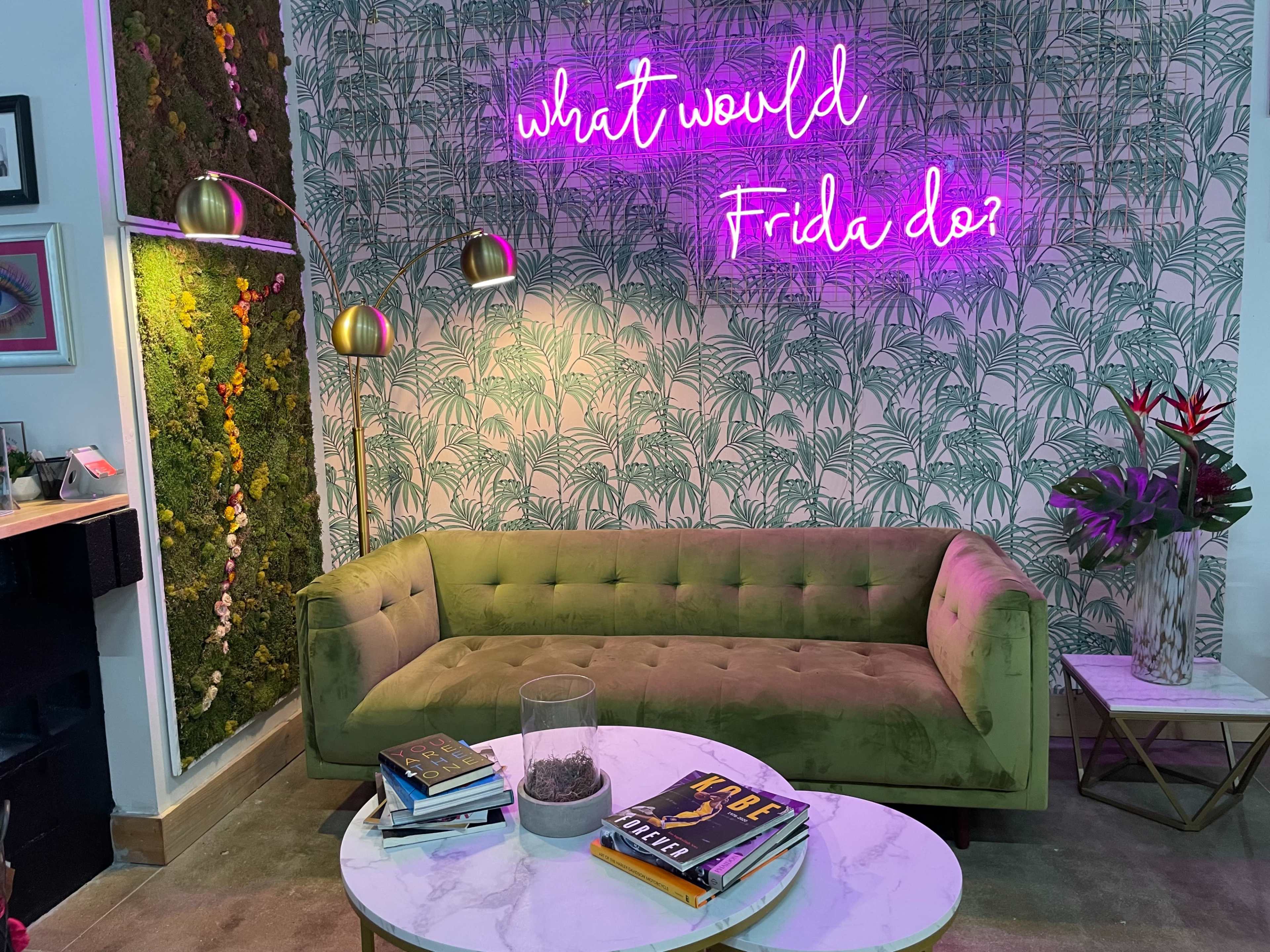 A green velvet sofa sits in front of a botanical-themed wall with a neon sign that reads, "what would Frida do?" and a coffee table adorned with books.