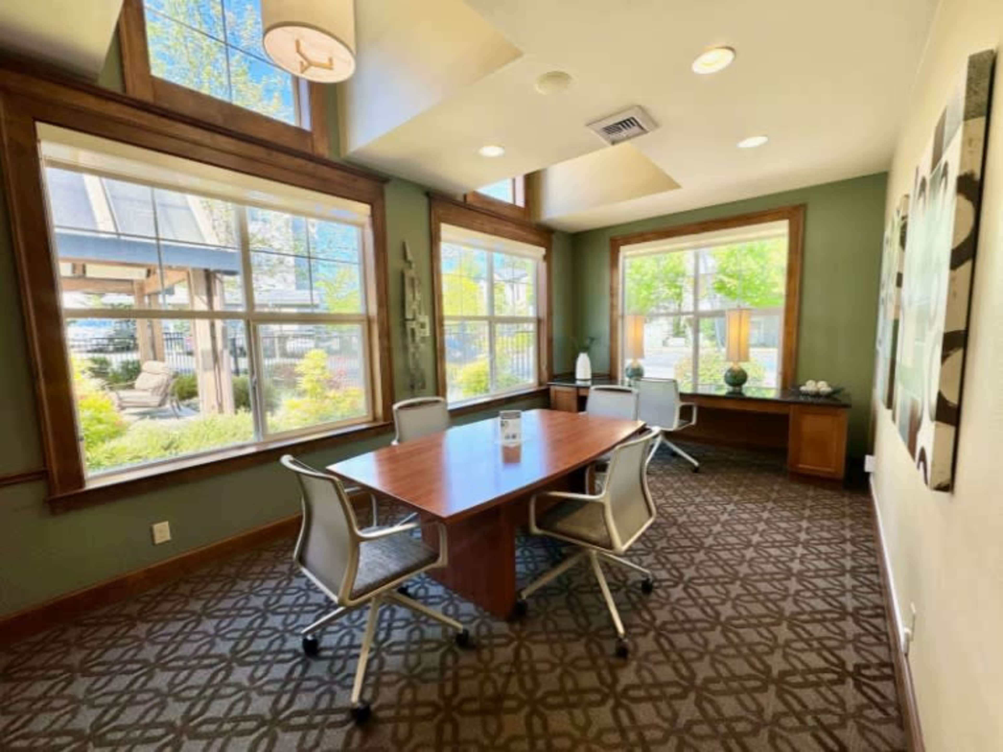 A small, well-lit conference room with a wooden table and four modern chairs, featuring large windows that provide views of greenery outside.