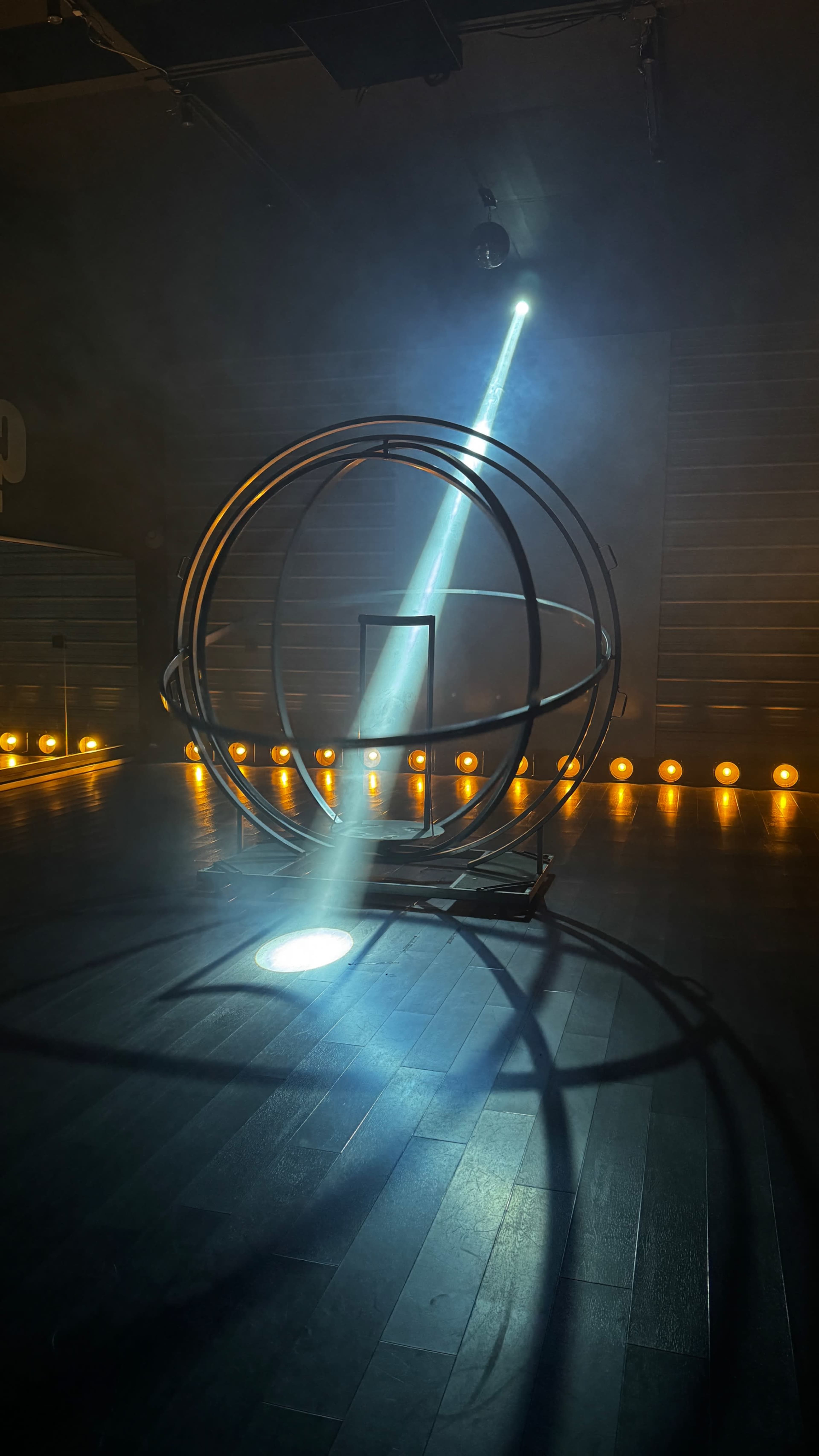 A circular metal structure is illuminated by a beam of light in a dimly lit room, with glowing lights lining the walls.
