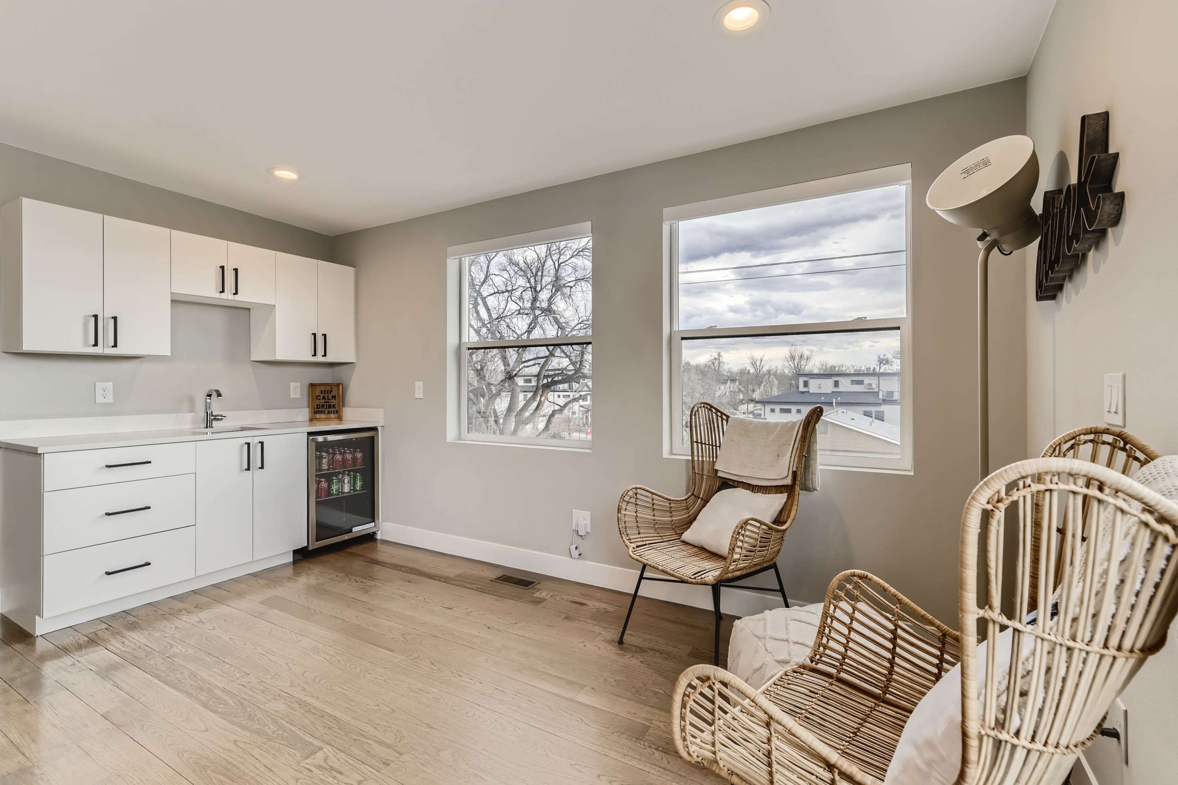 A bright, modern room features a kitchenette with white cabinets, two large windows, and two wicker chairs.