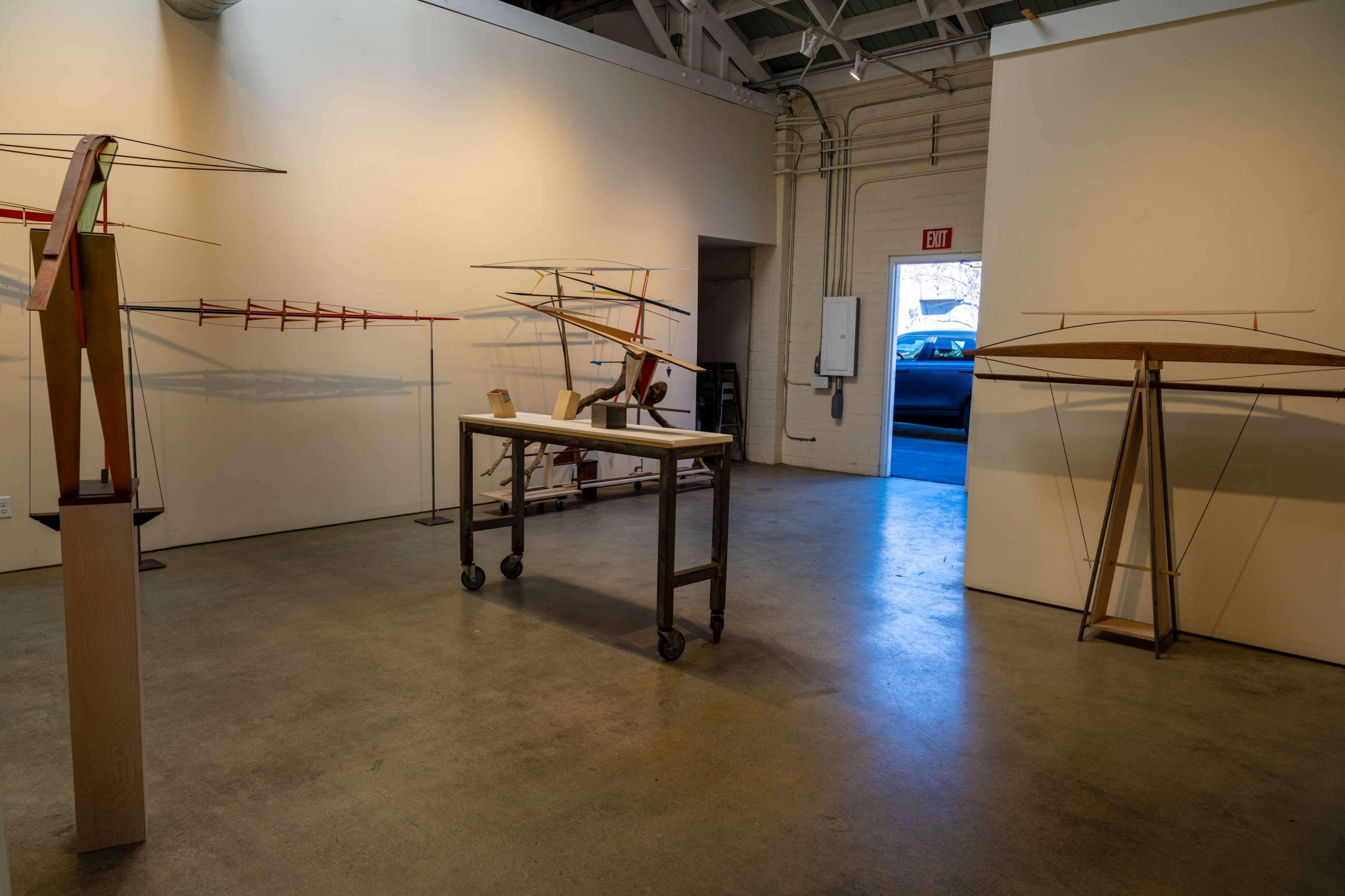 The image shows an art gallery space featuring several wooden models of flying machines displayed on pedestals and a table, with a doorway leading outside.