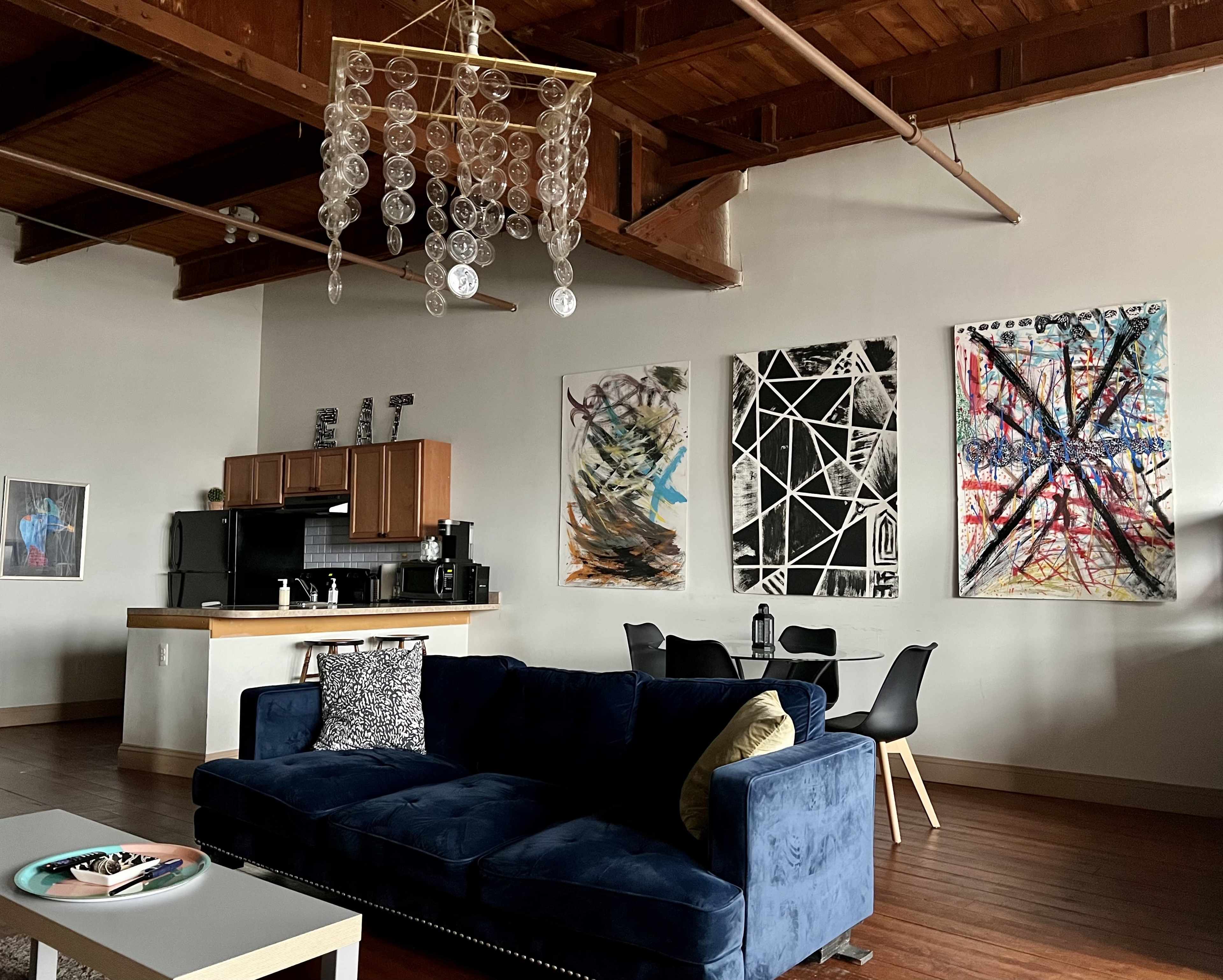 A spacious living area with a blue couch, a modern dining table and chairs, artistic wall hangings, and a kitchen area in the background.