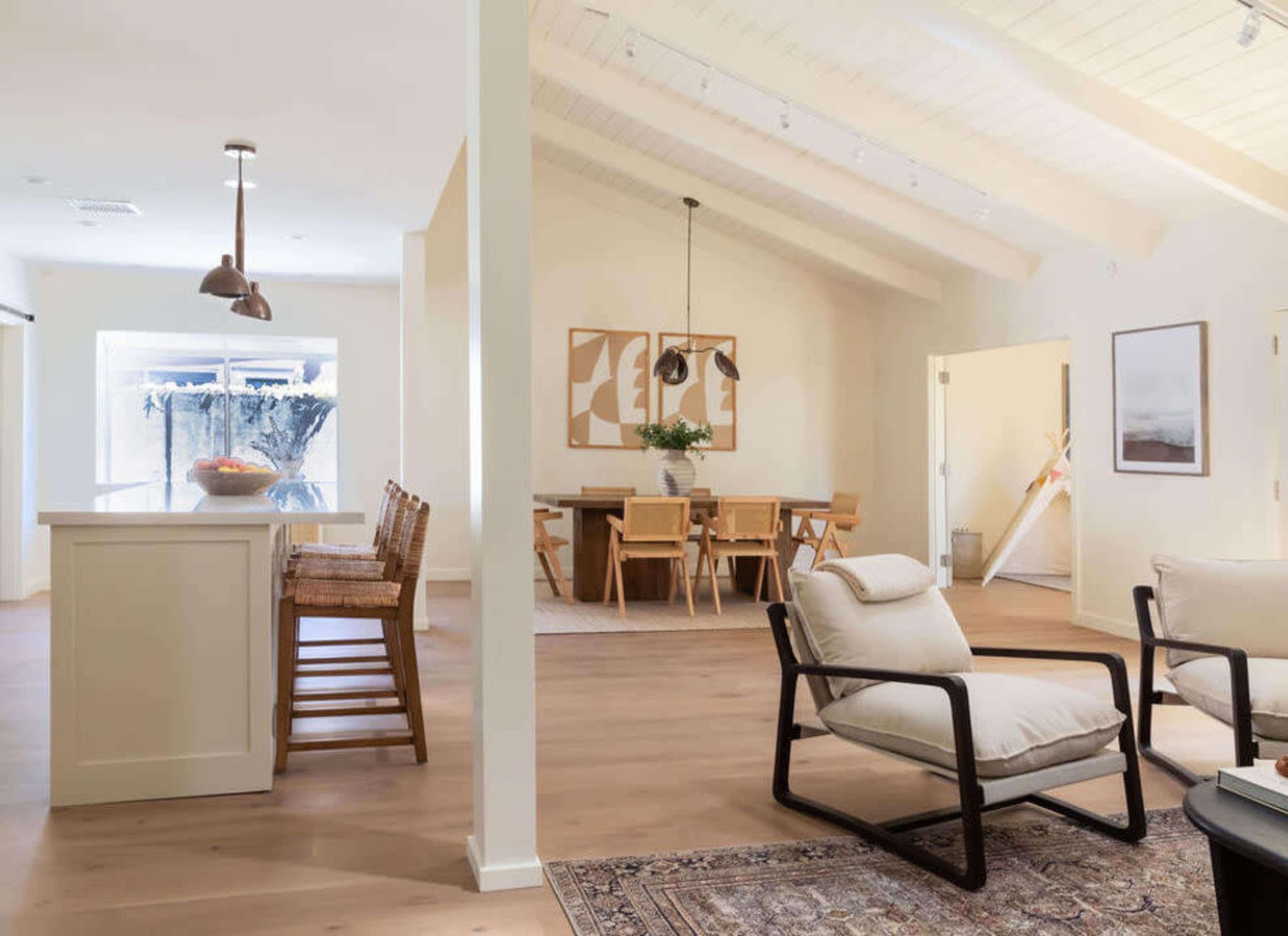 The image shows a modern open-concept living space featuring a kitchen with bar stools, a dining area with wooden chairs, and a cozy seating arrangement with a rug.