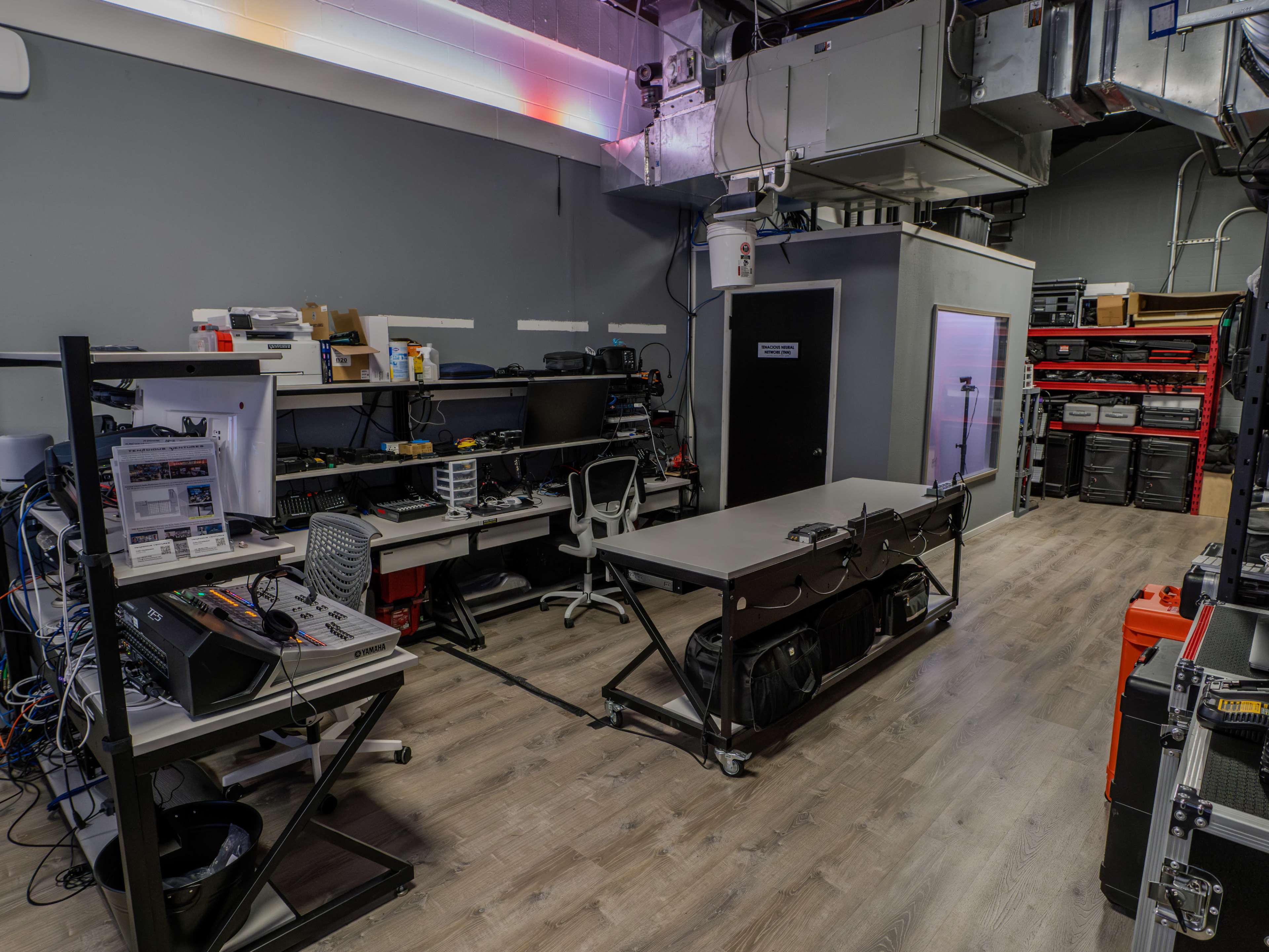 The image shows a spacious workspace with desks, equipment, and storage areas, featuring various tools and electronic devices organized along the walls.