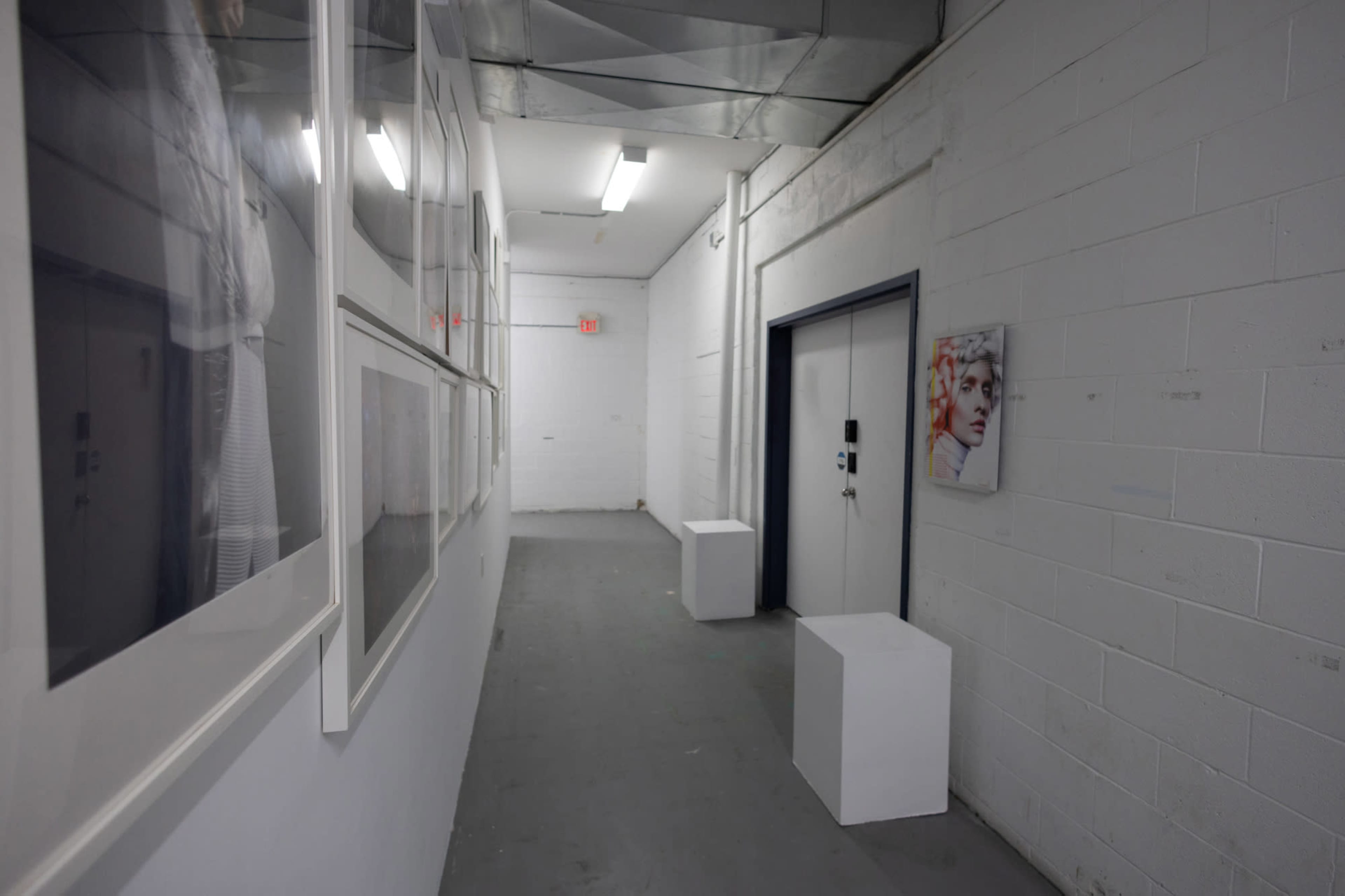 A narrow hallway with white walls features framed photographs on one side and a door with a small portrait hanging adjacent to it.