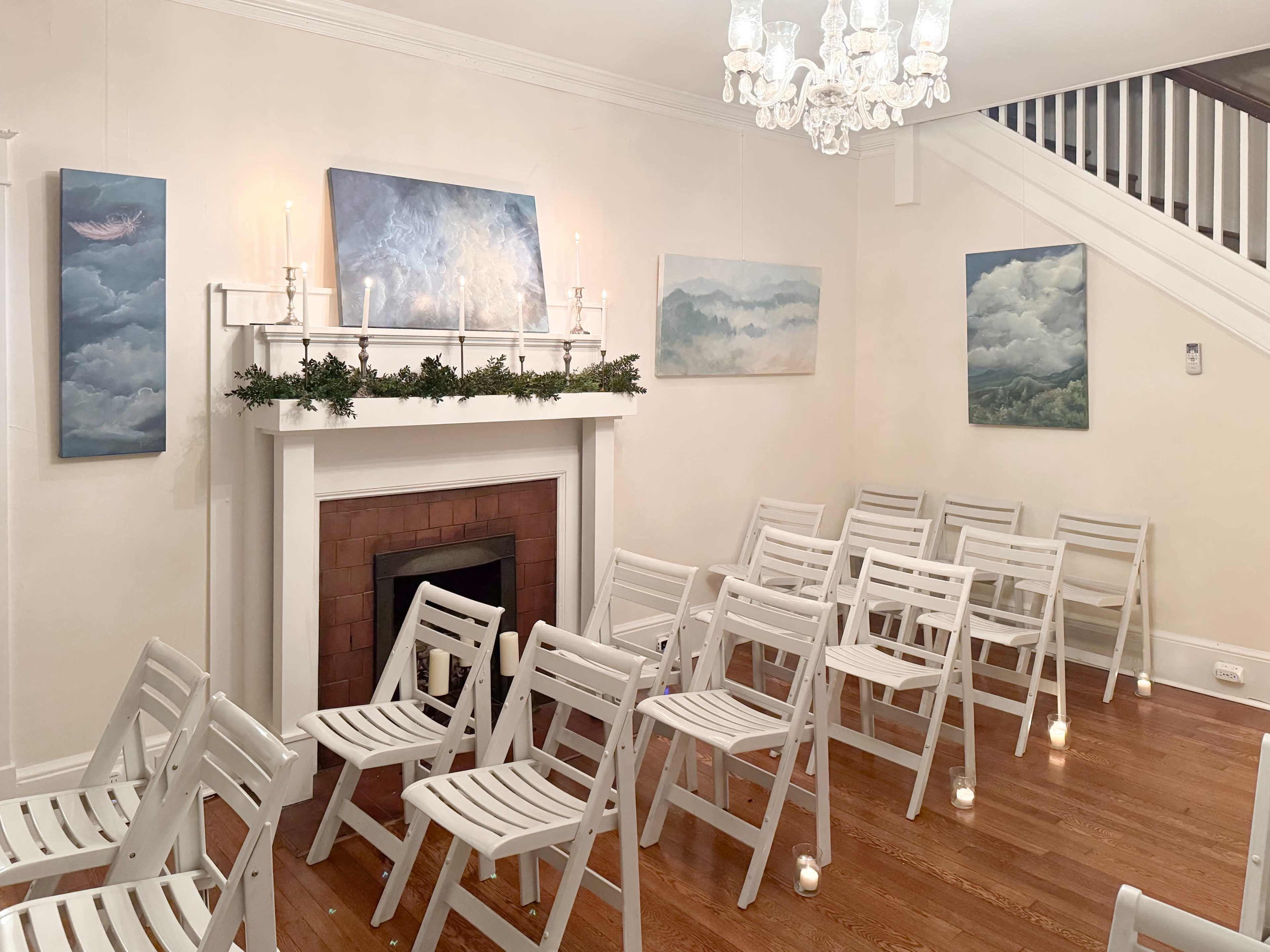 A room with white folding chairs arranged in front of a fireplace, decorated with artwork and greenery, and illuminated by a chandelier.