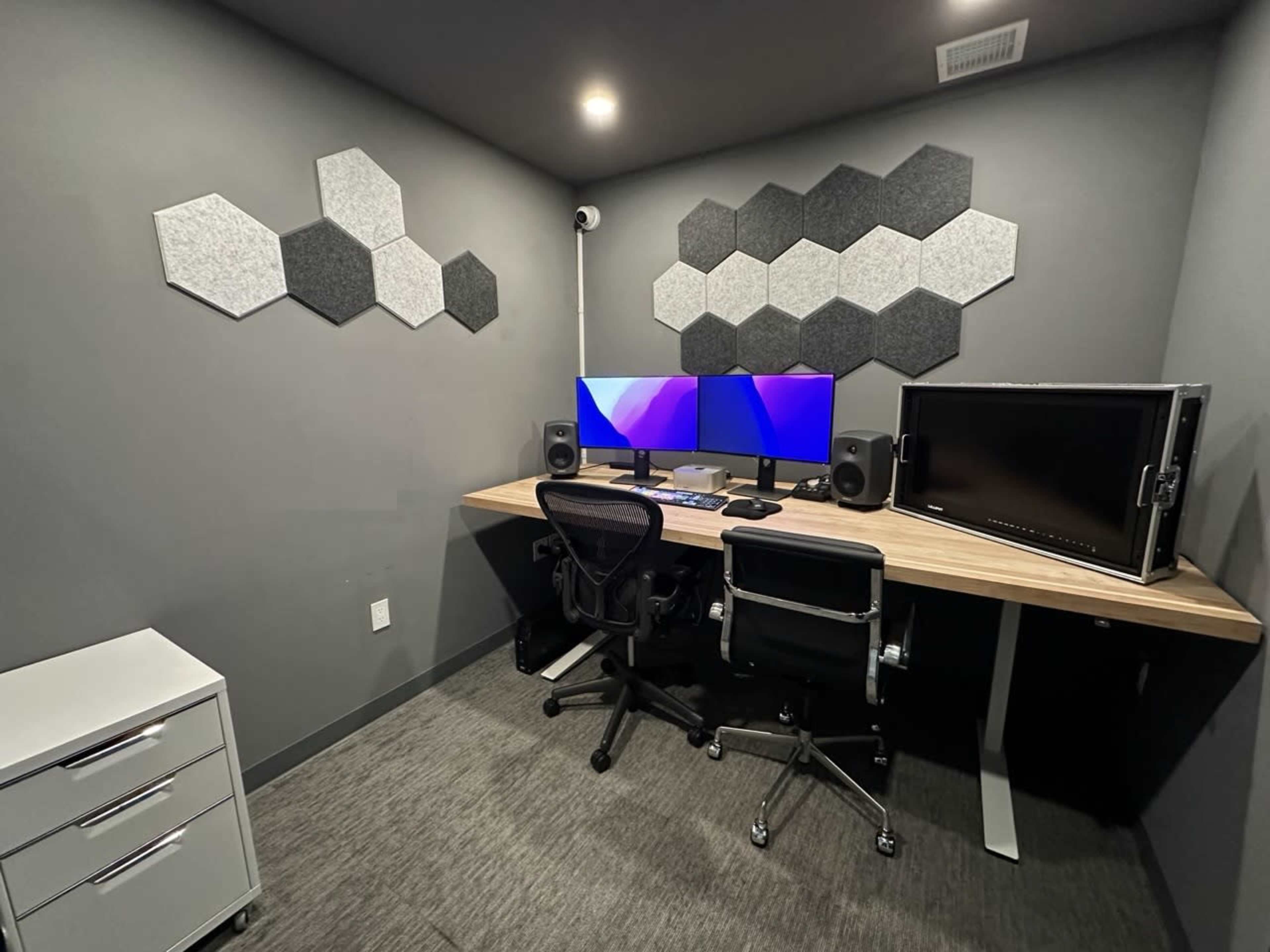 A modern workspace features two black office chairs, a wooden desk with two computer monitors, and hexagon-shaped acoustic panels on the gray walls.