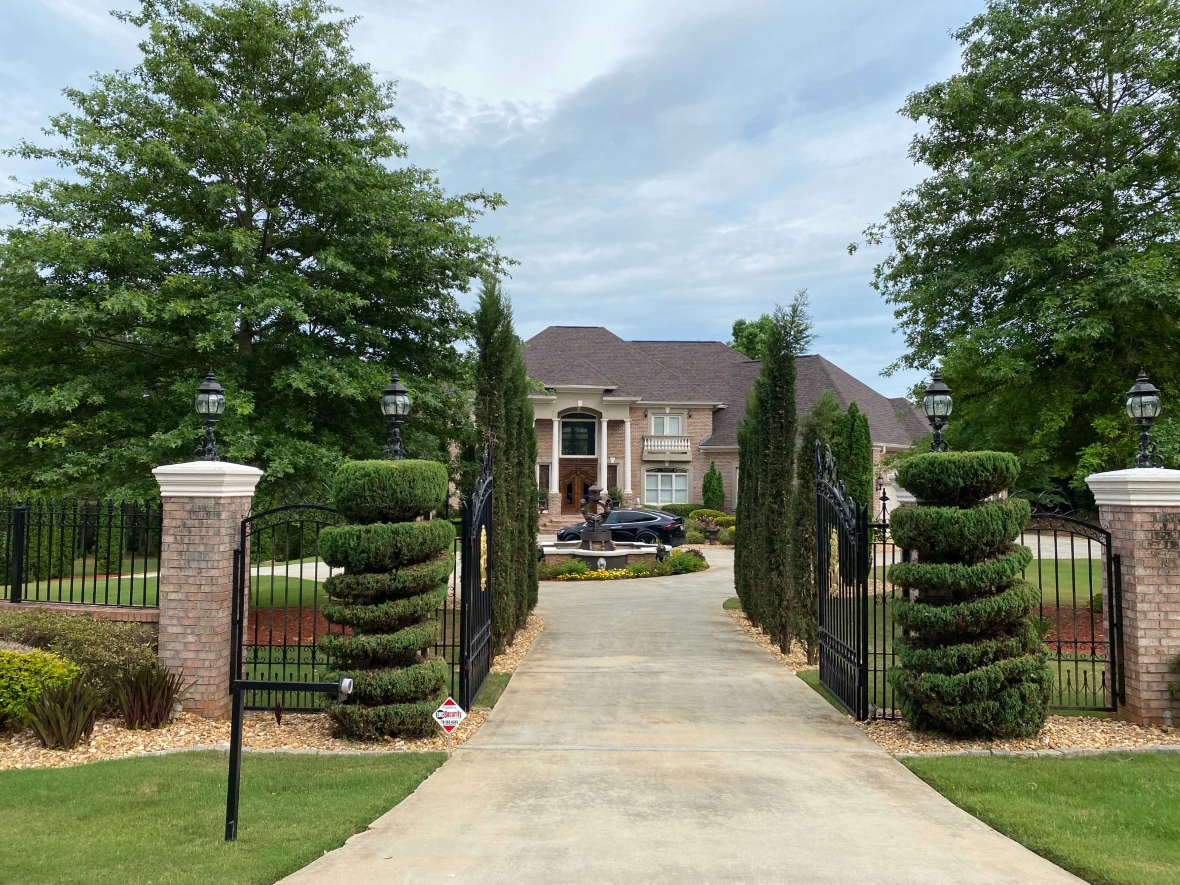 A paved driveway leads to a large house surrounded by manicured hedges and trees, with an iron gate at the entrance.