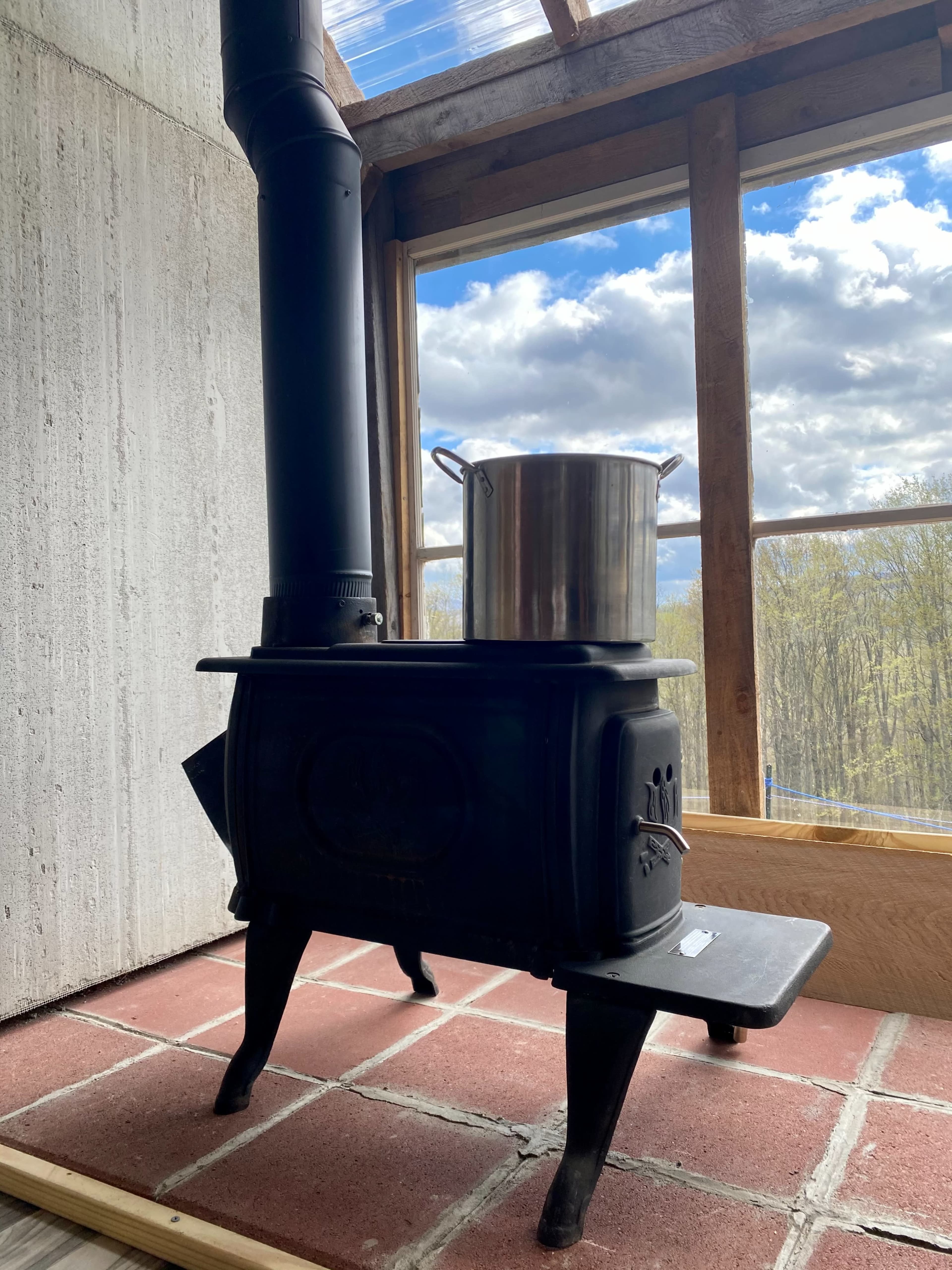 A metal stove with a pot on top stands on a brick floor beside a window with a view of trees and clouds.