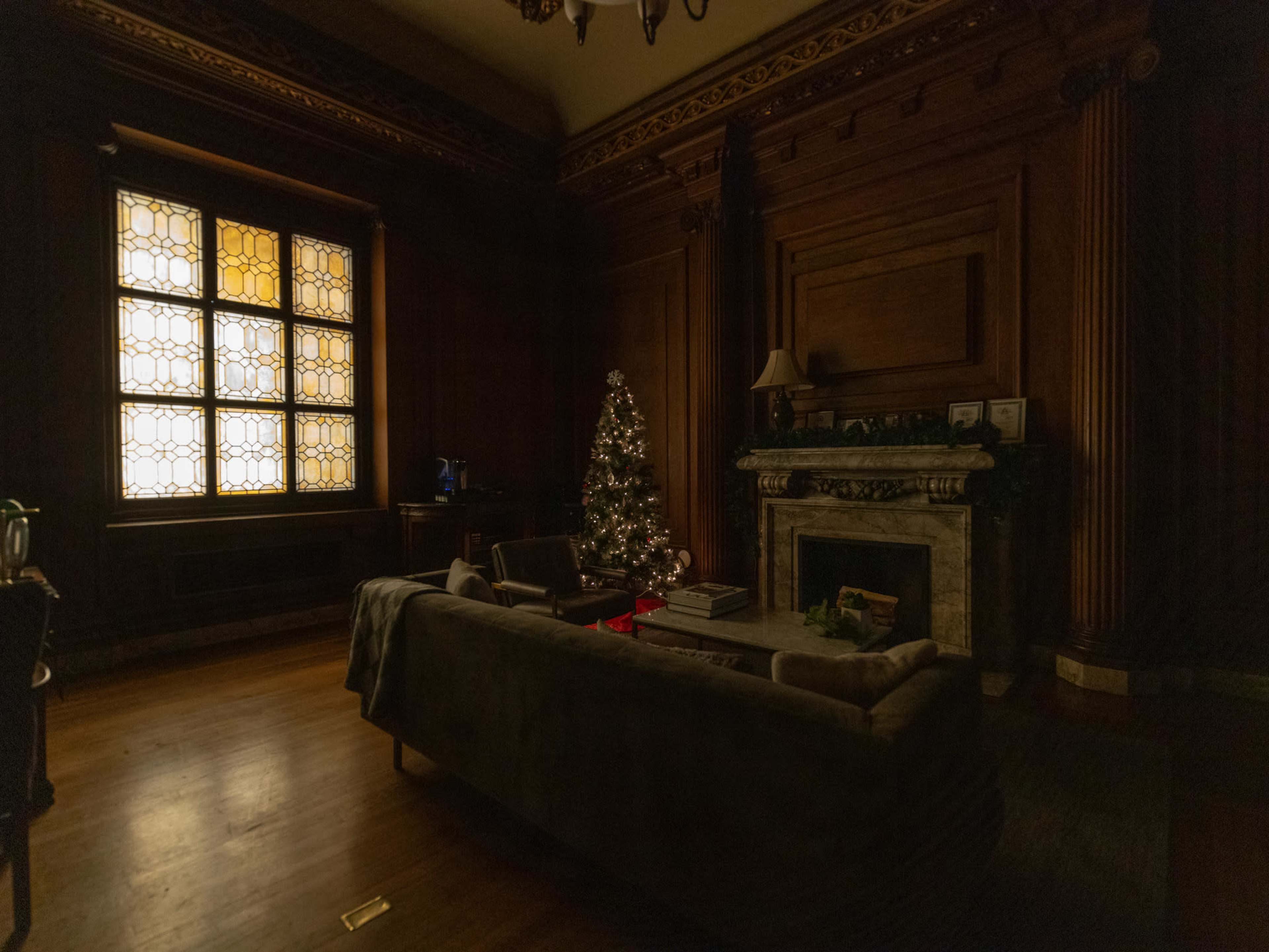 A dimly lit living room features a Christmas tree beside a fireplace, with ornate wooden walls and a large stained glass window.