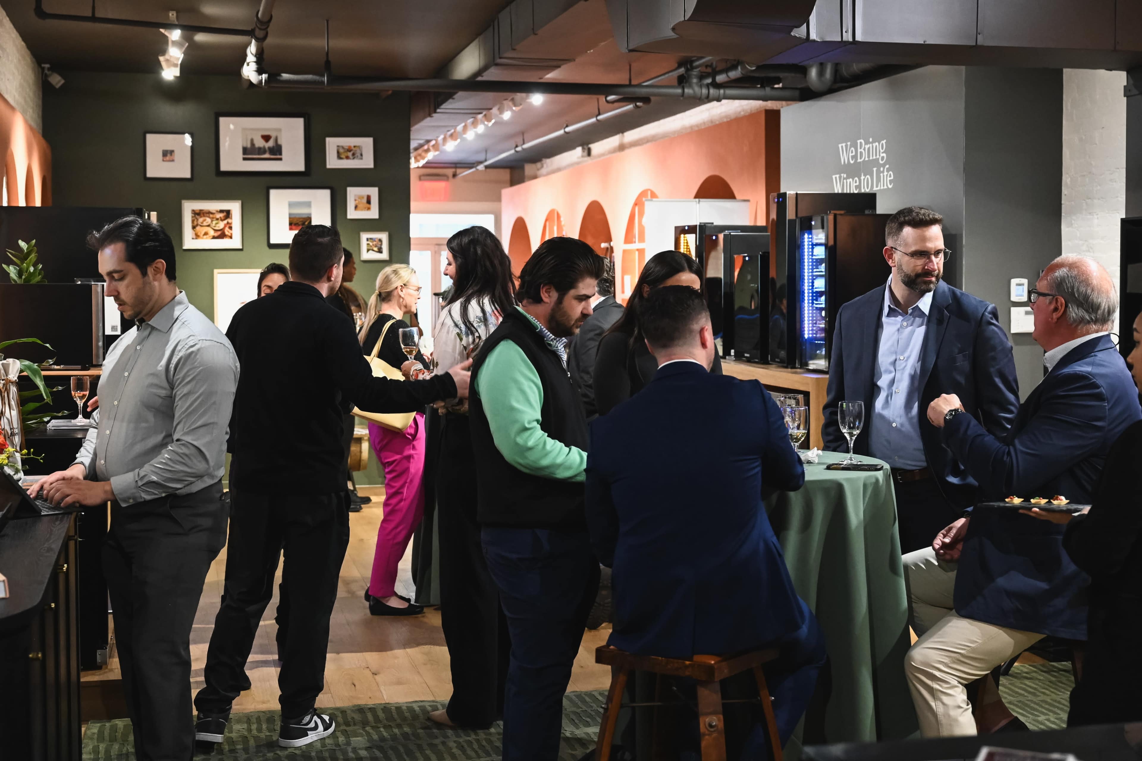 A group of people mingles at a social event in a well-decorated interior space, with some engaged in conversation and others enjoying drinks and food.