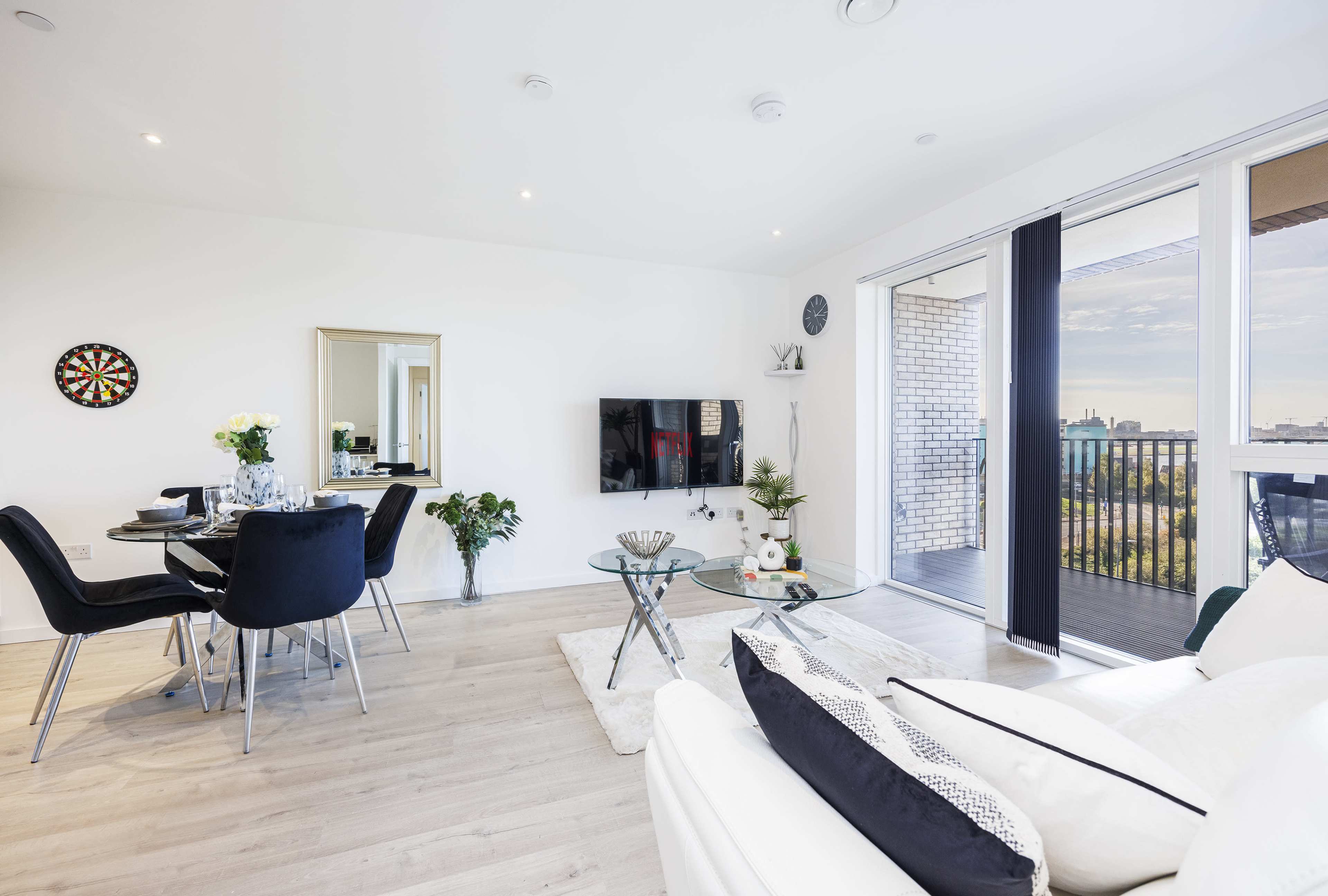 A modern living room with a dining area, featuring a glass table, black chairs, a white sofa, a television, and a balcony with large glass doors.