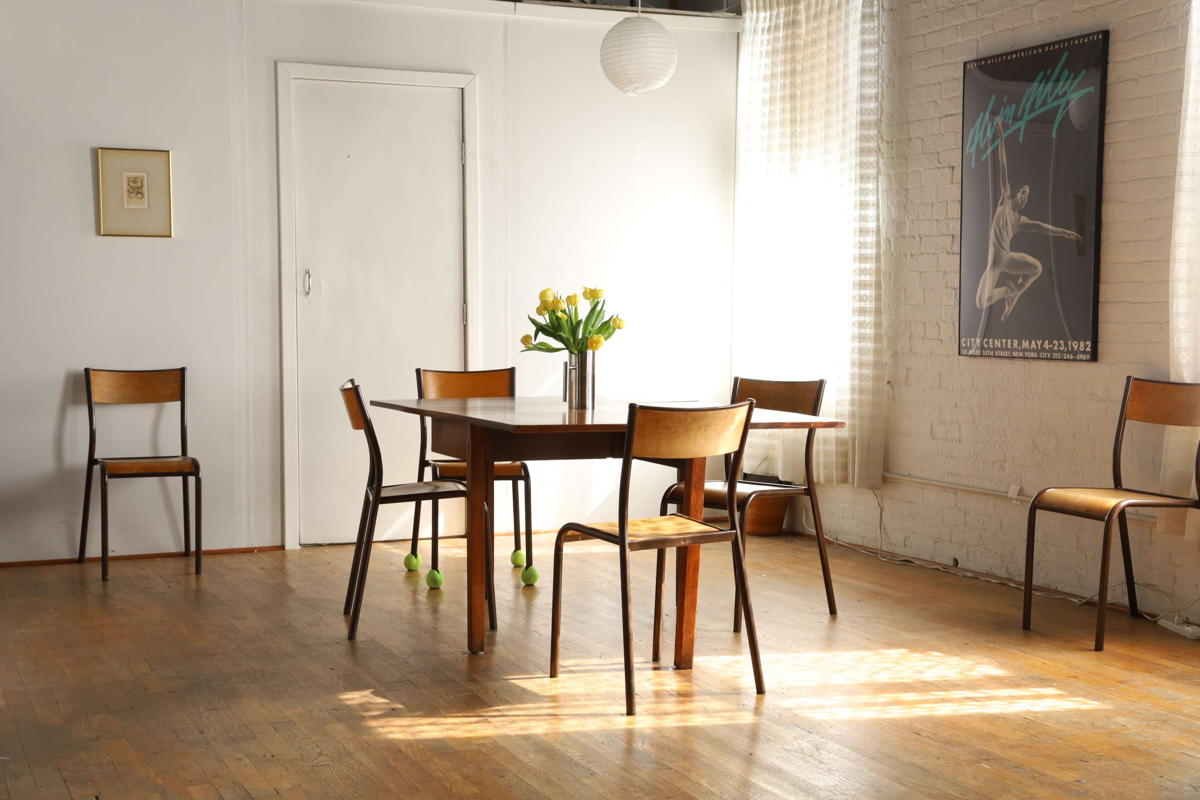 A wooden table surrounded by chairs is positioned in a sunlit room with a vase of yellow flowers and a poster on the wall.