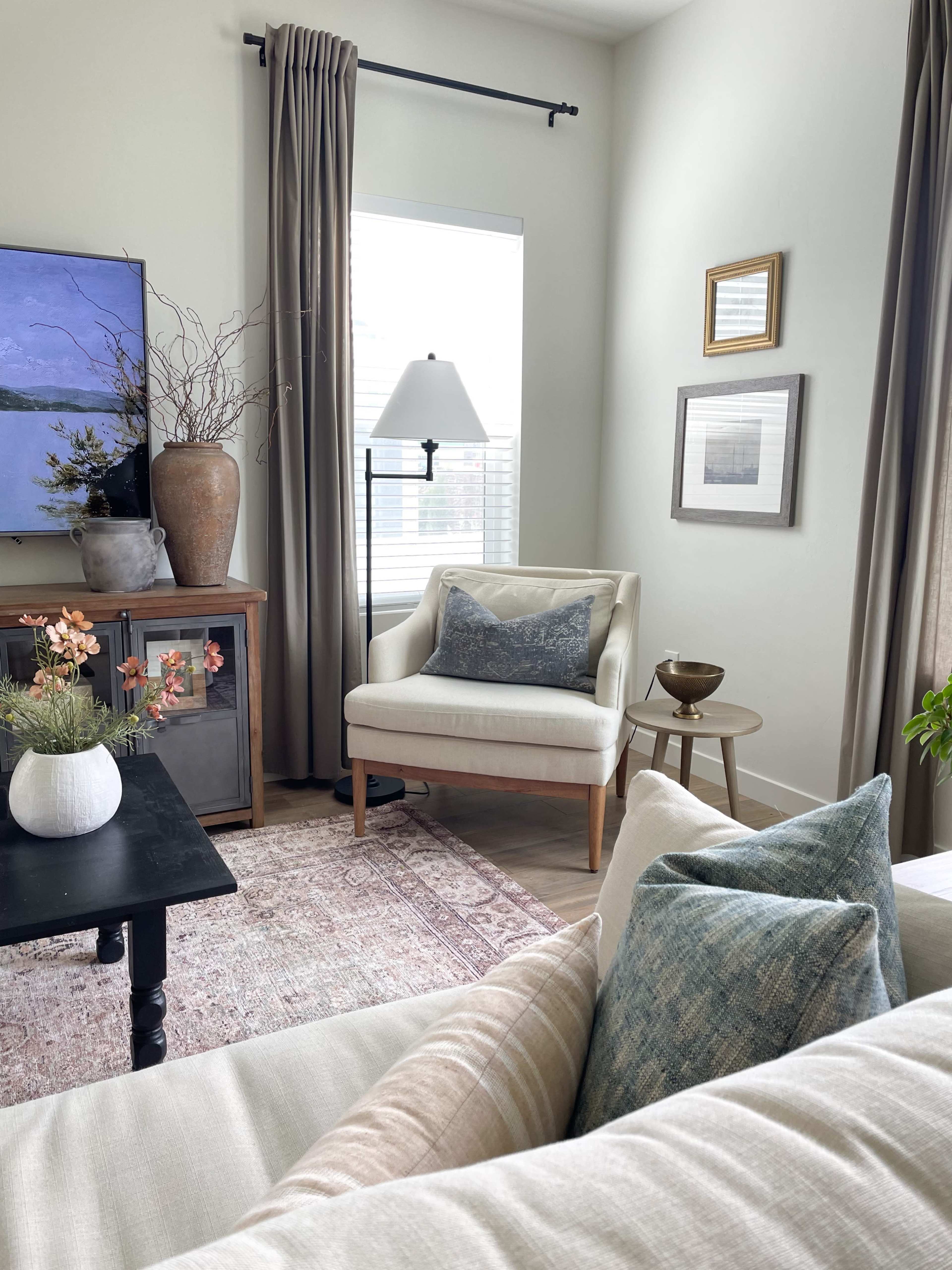 A living room features a beige armchair next to a wooden side table, with a gray couch, decorative pillows, and a large window adorned with curtains.