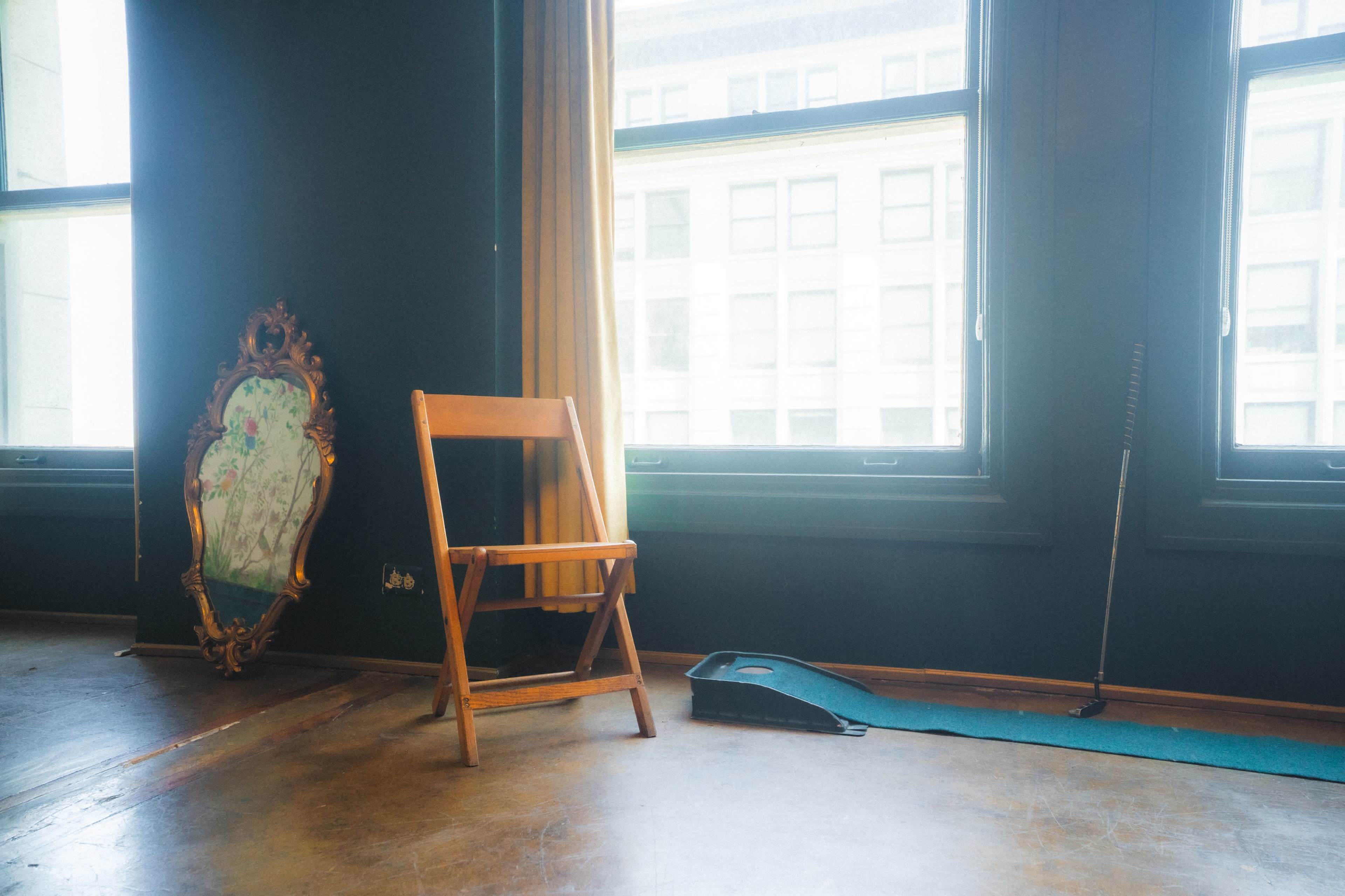 The image shows a wooden folding chair near a mirror and a small putting green in a brightly lit room with large windows.
