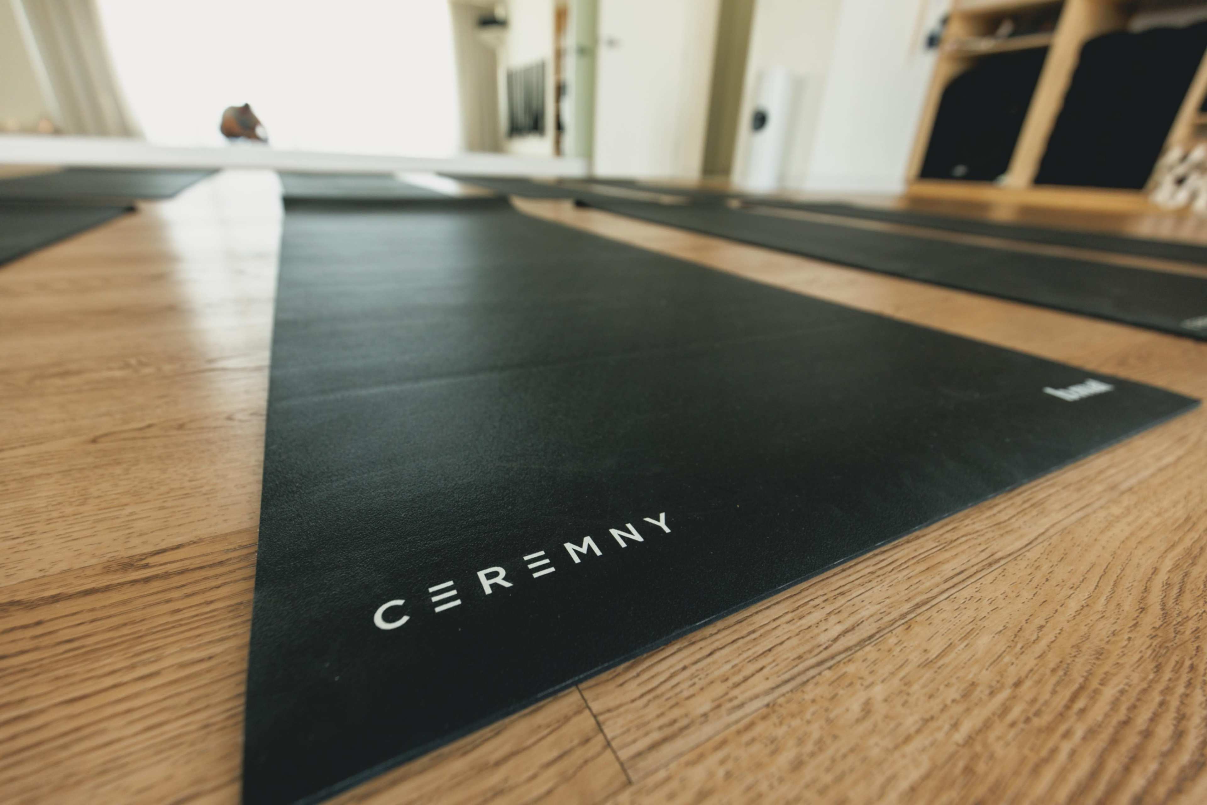 The image shows a close-up view of black yoga mats with the word "CEREMNY" printed on them, laid out on a wooden floor in a well-lit space.