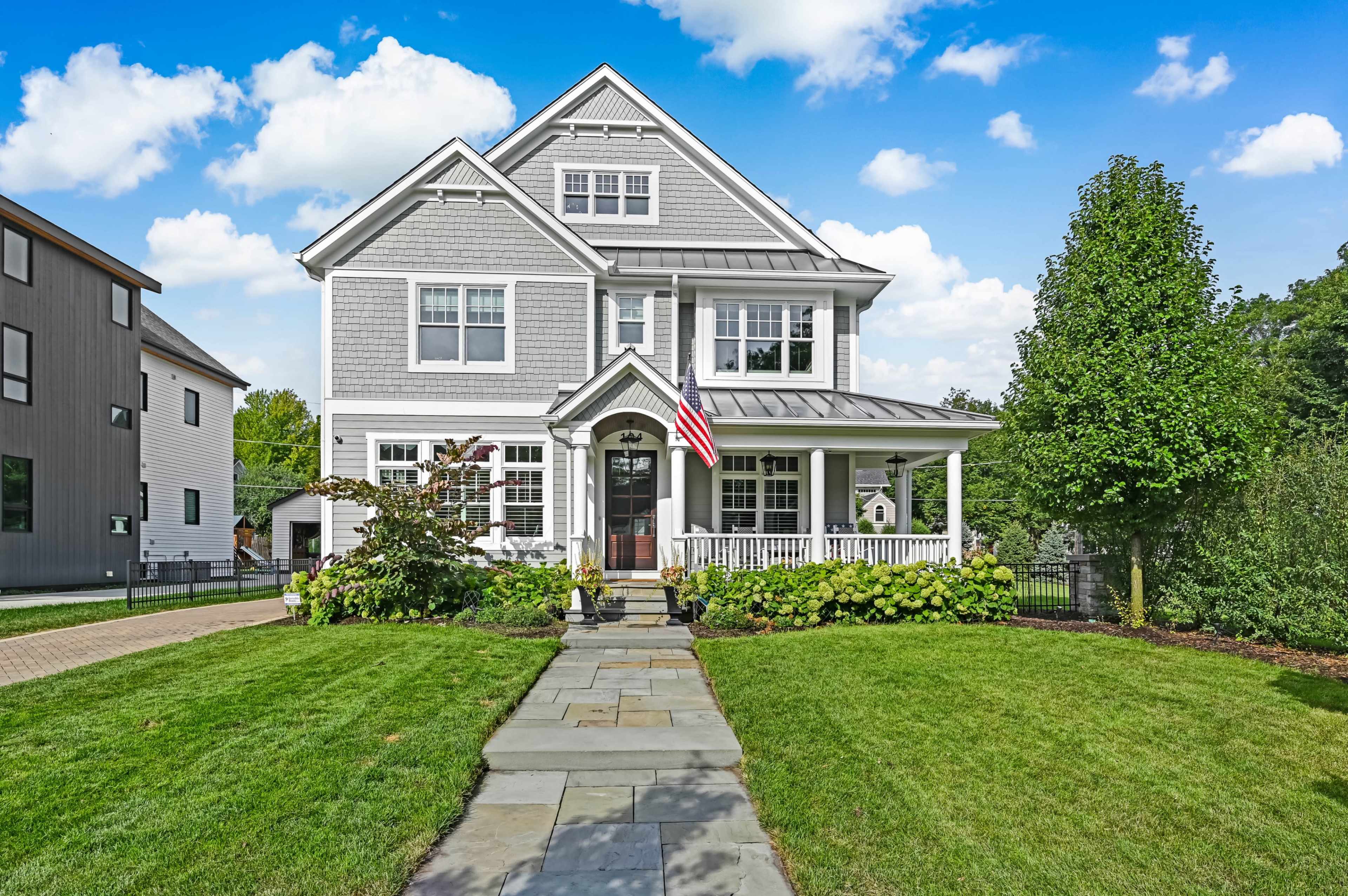 The image shows a large, multi-story gray house with a front porch and an American flag, surrounded by a well-maintained garden and lawn.