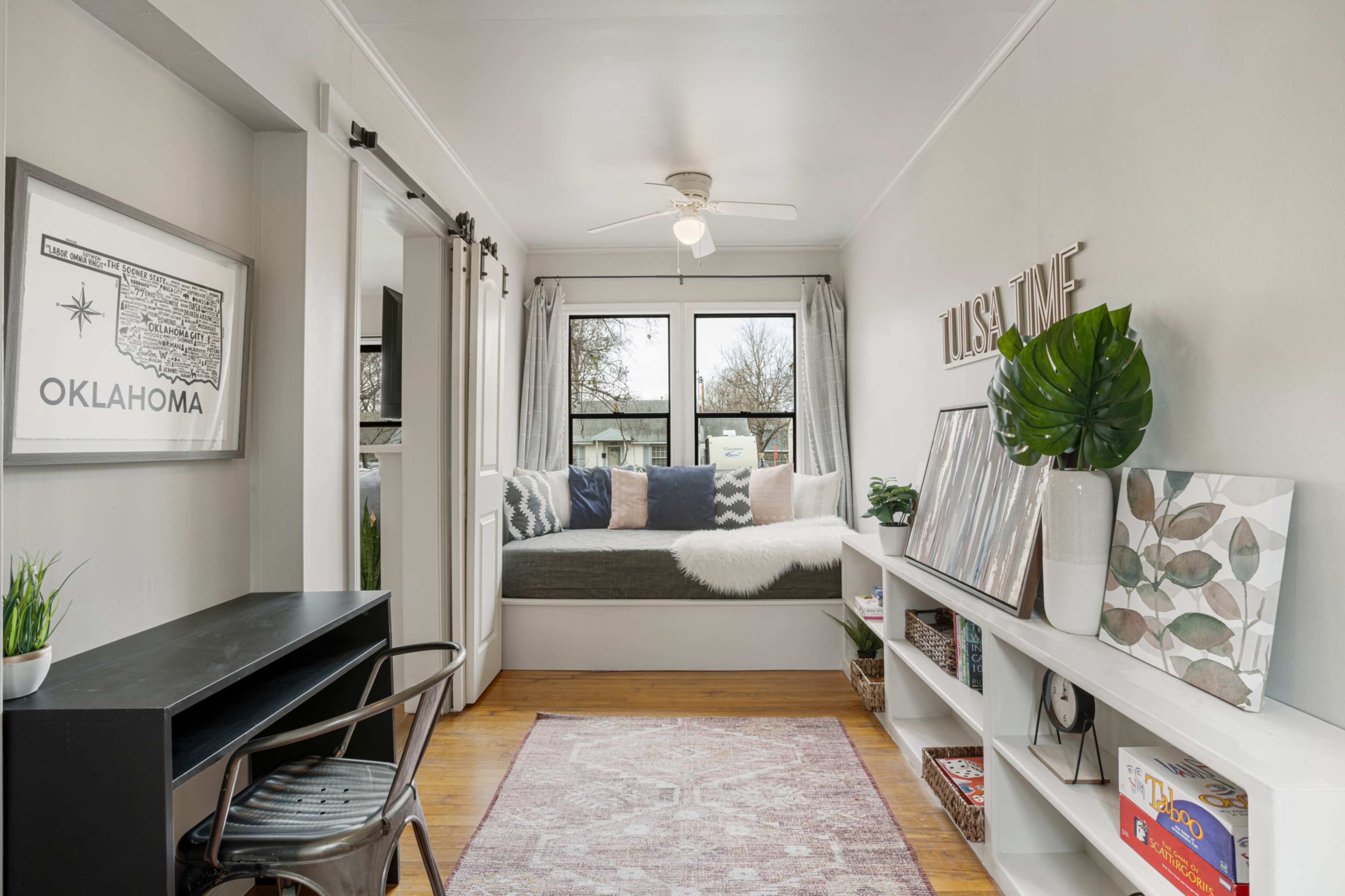 A cozy room features a window seat with cushions, a desk on the left, and a shelf displaying decorative items and plants.