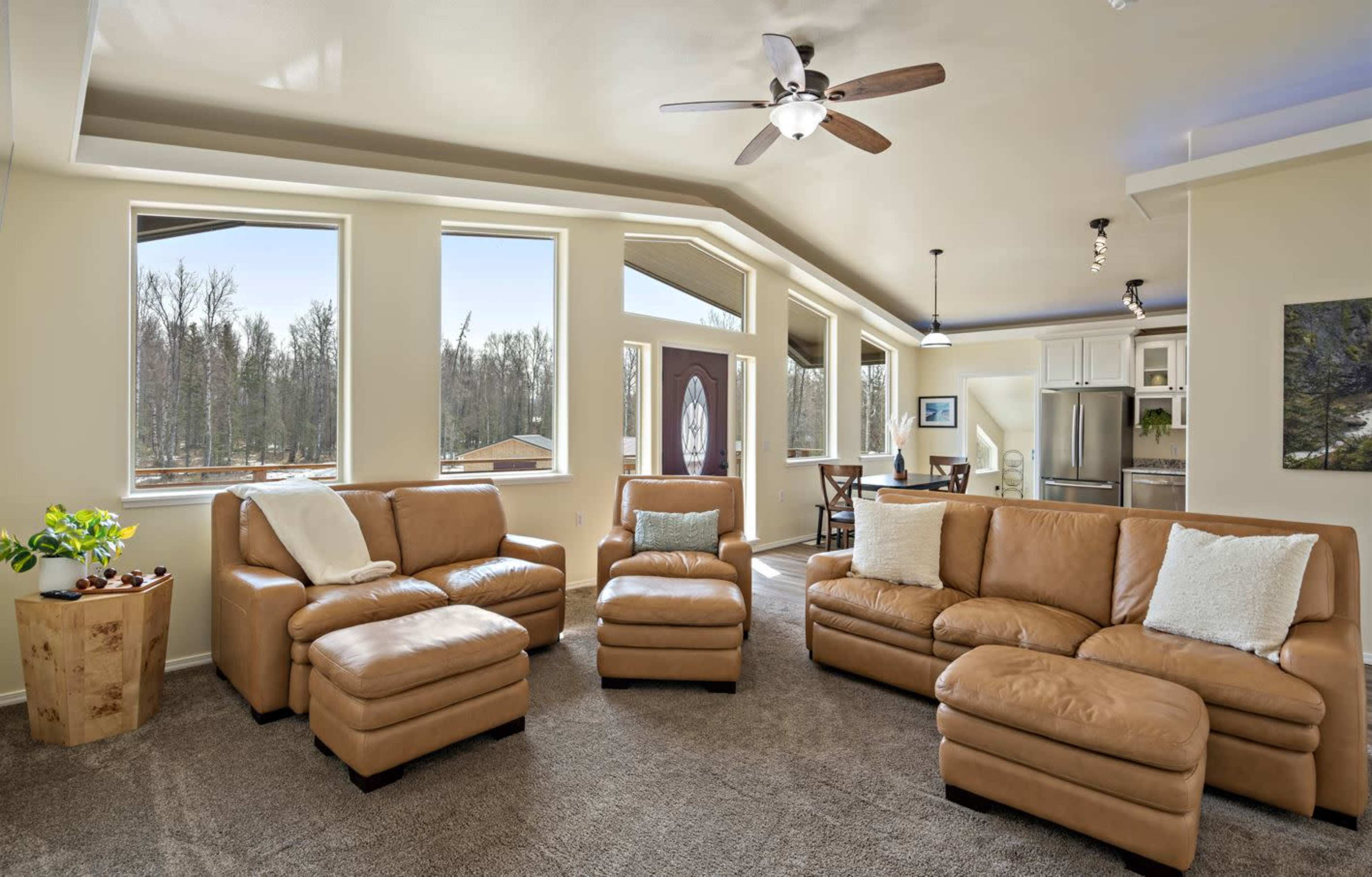 A modern living room with light-colored walls, a sectional leather couch, and large windows providing a view of a forested area outside.