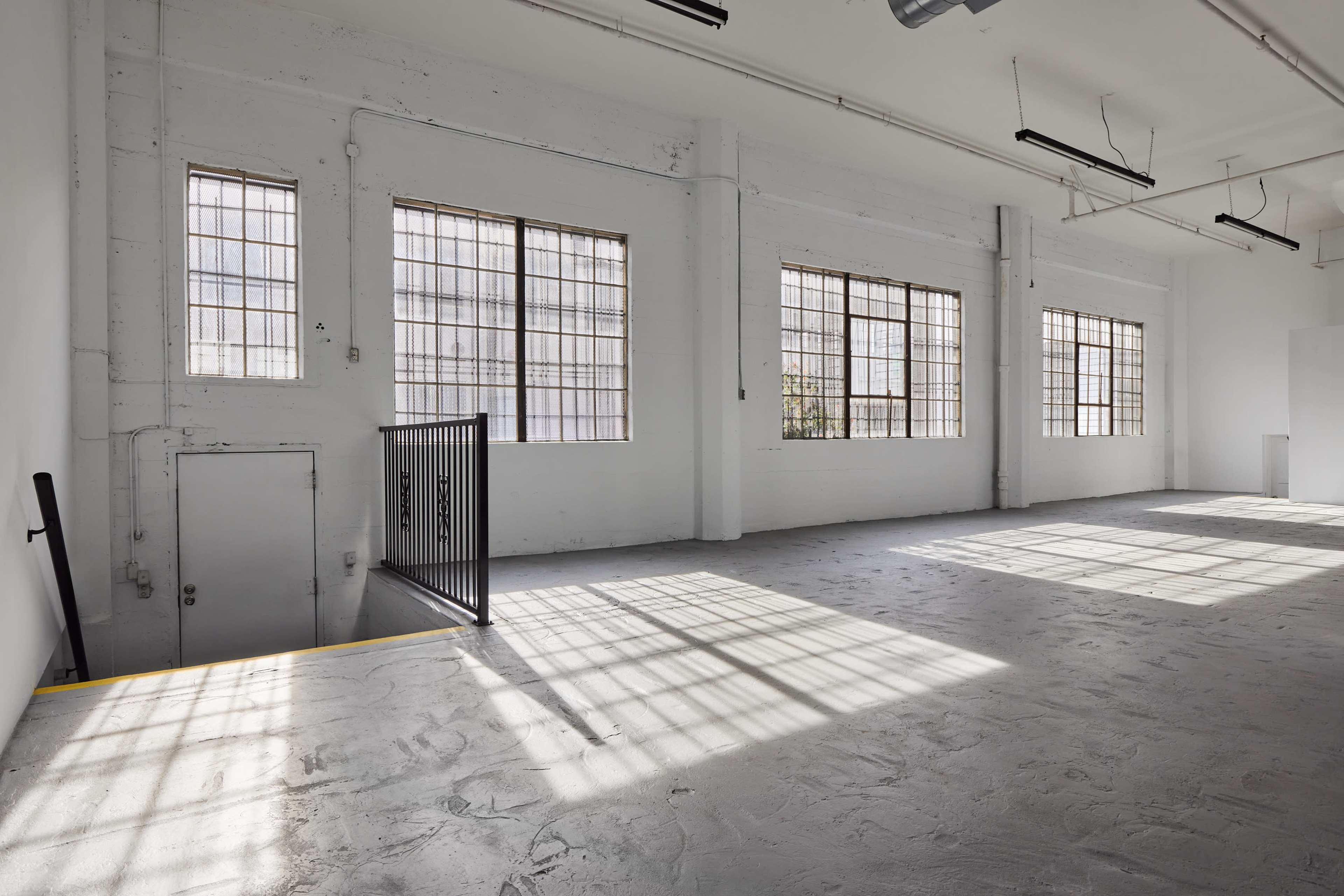 The image shows a spacious, minimally furnished room with large windows allowing natural light to create shadow patterns on the concrete floor.