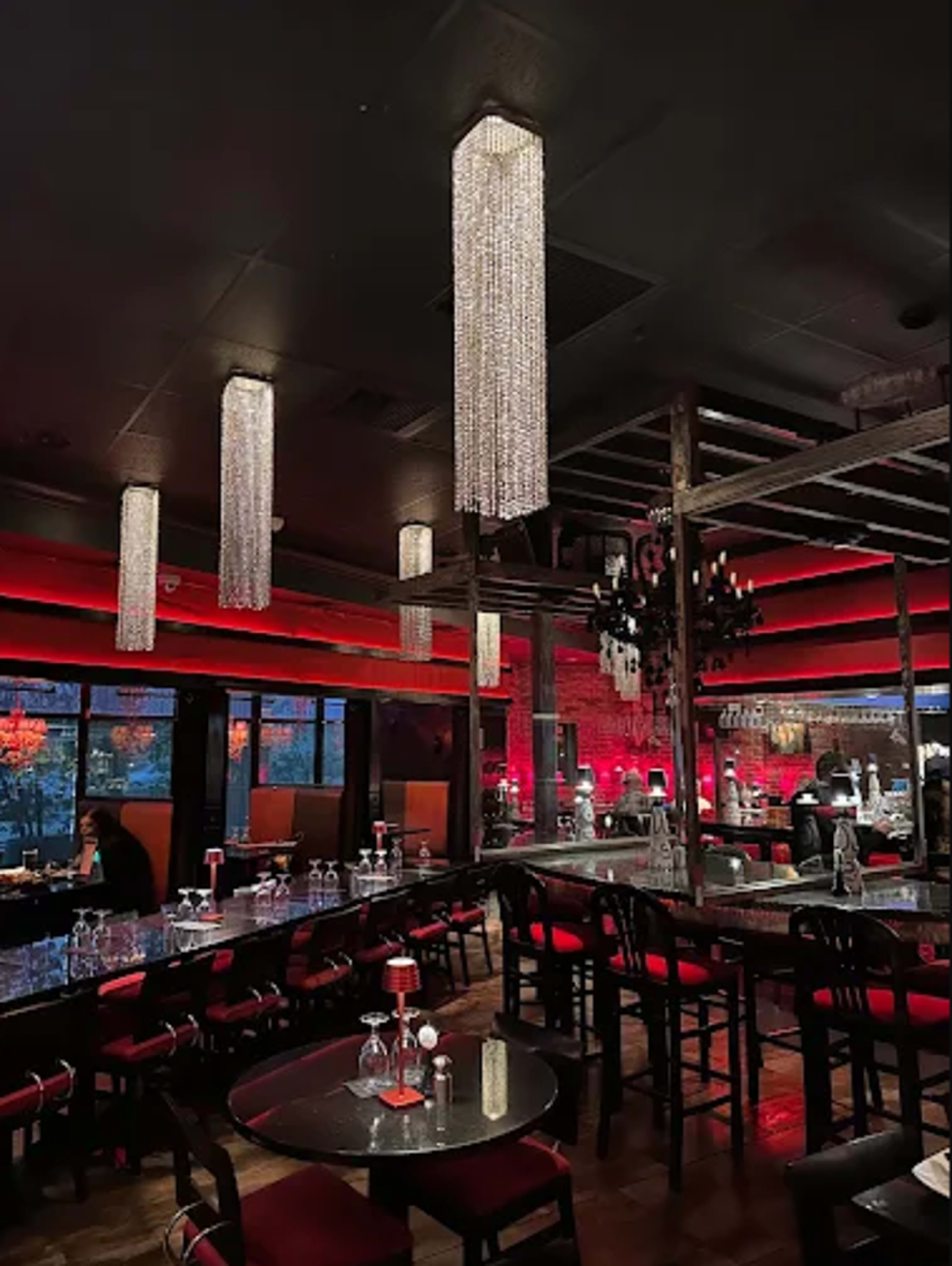 The interior of a dimly lit restaurant features several crystal chandeliers hanging from the ceiling, red accents, and black furniture with red upholstery.