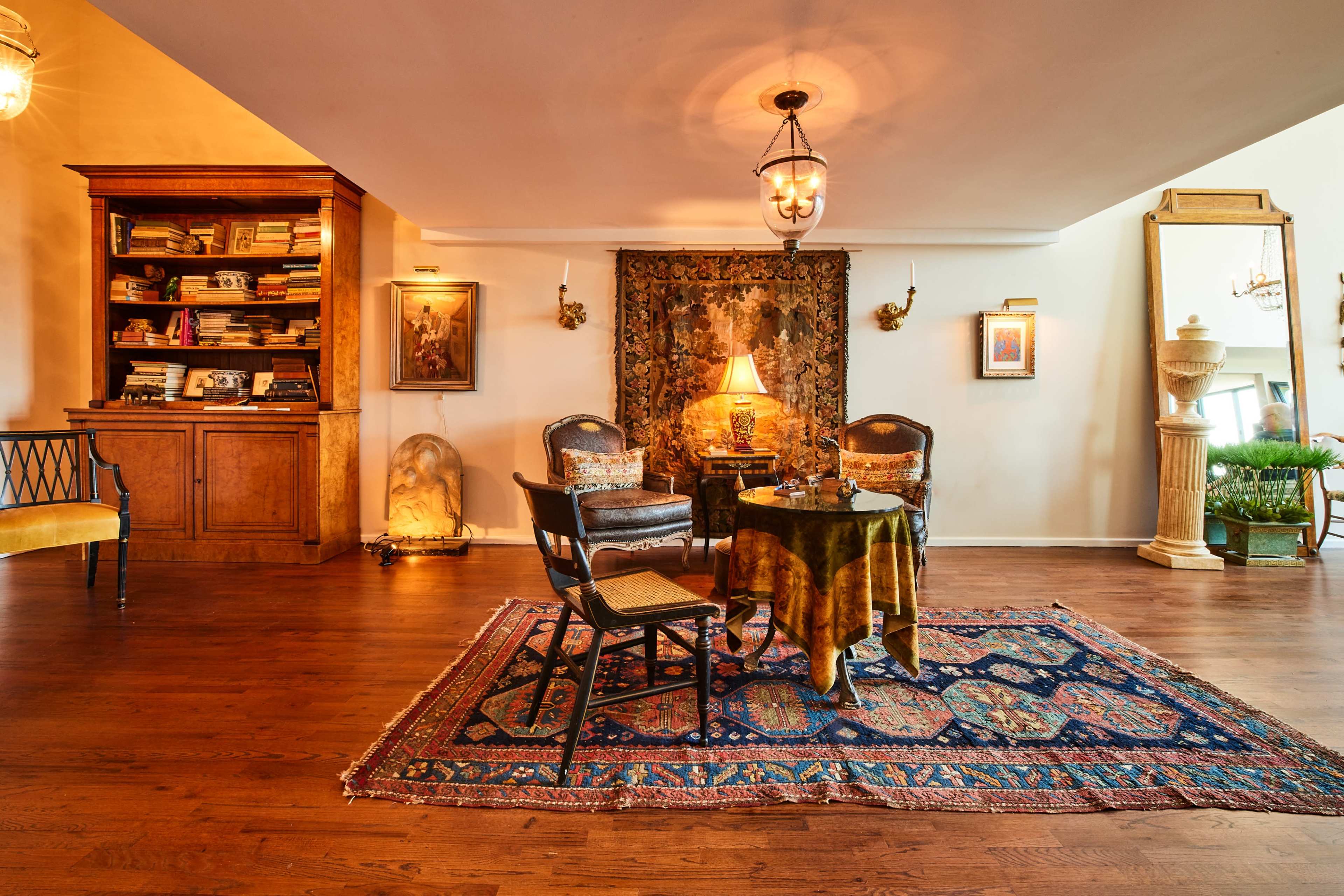 The image shows a cozy room with hardwood floors, featuring a vintage area rug, a small table with chairs, a bookshelf filled with books, and decorative artwork on the walls.