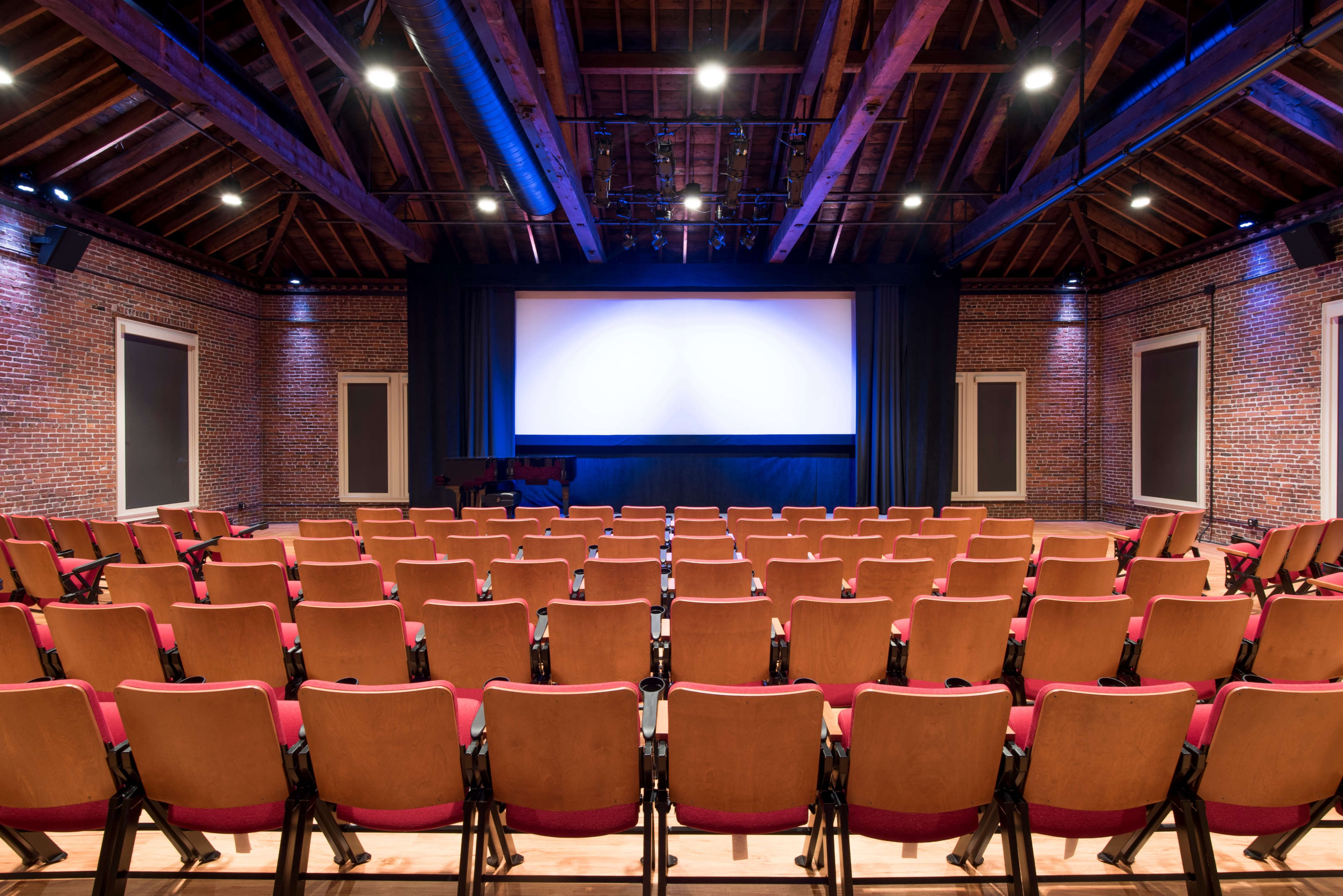 An empty auditorium with wooden beams, brick walls, and rows of seating facing a large screen.