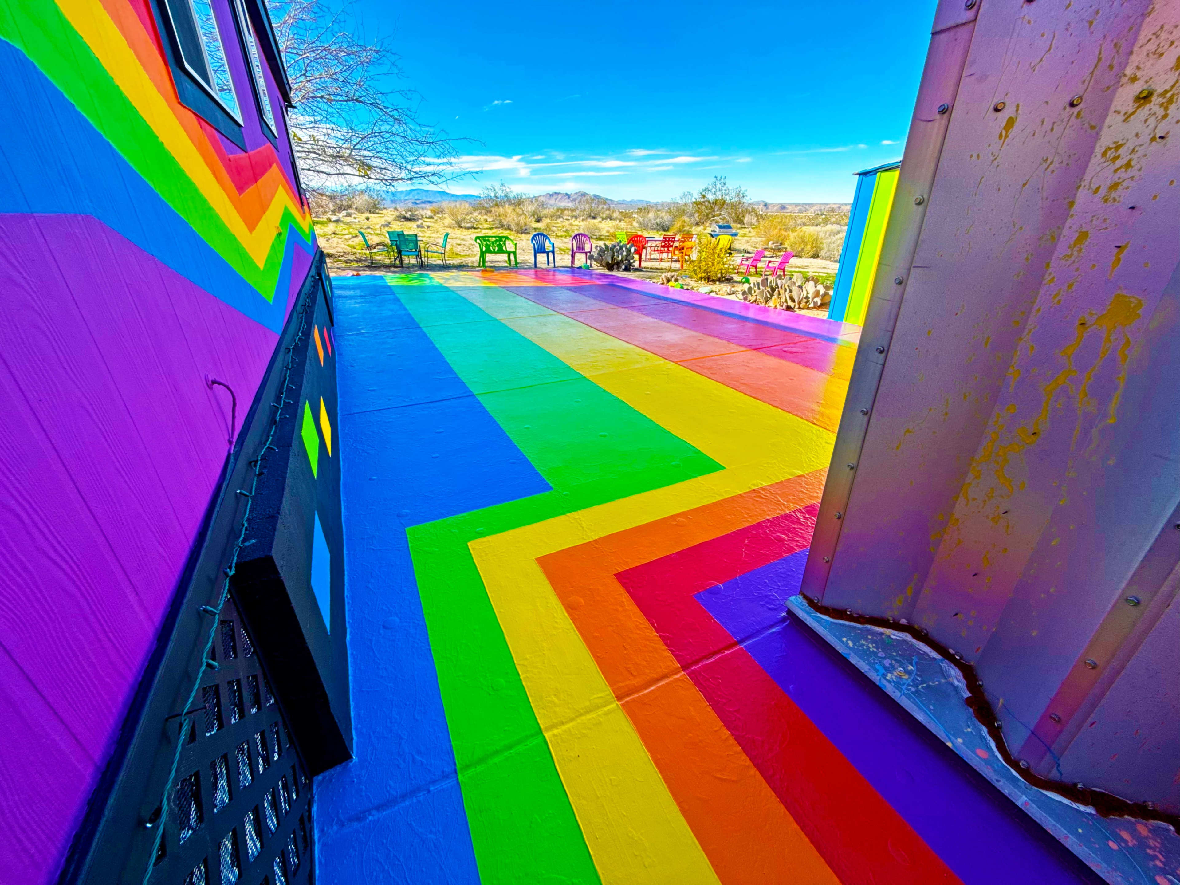 The image shows a vibrant, multi-colored rainbow pathway leading to a patio area, surrounded by colorful chairs and a clear blue sky.