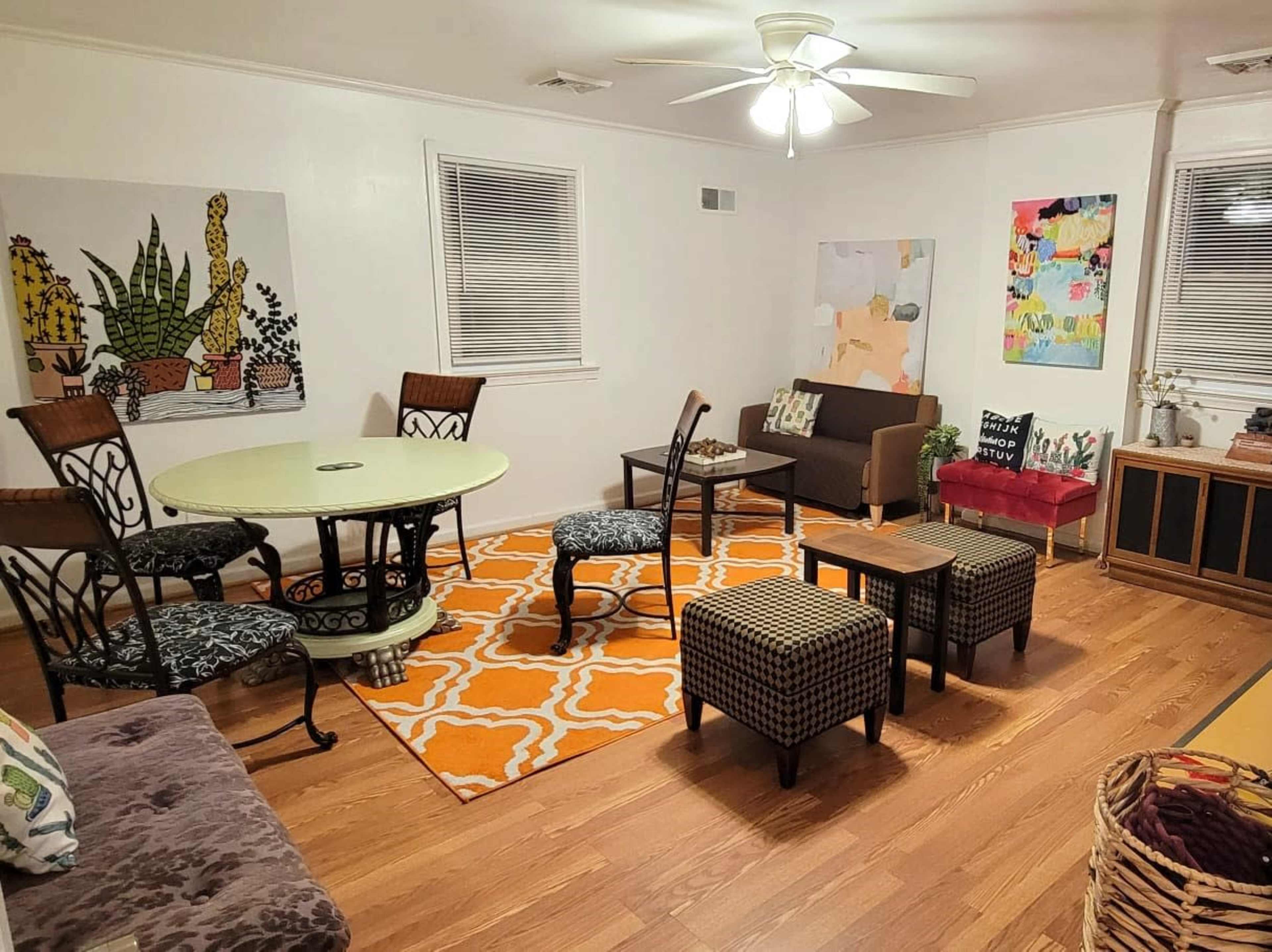 The room features a circular table surrounded by several chairs, with artwork on the walls and a mix of seating arrangements on a patterned rug.