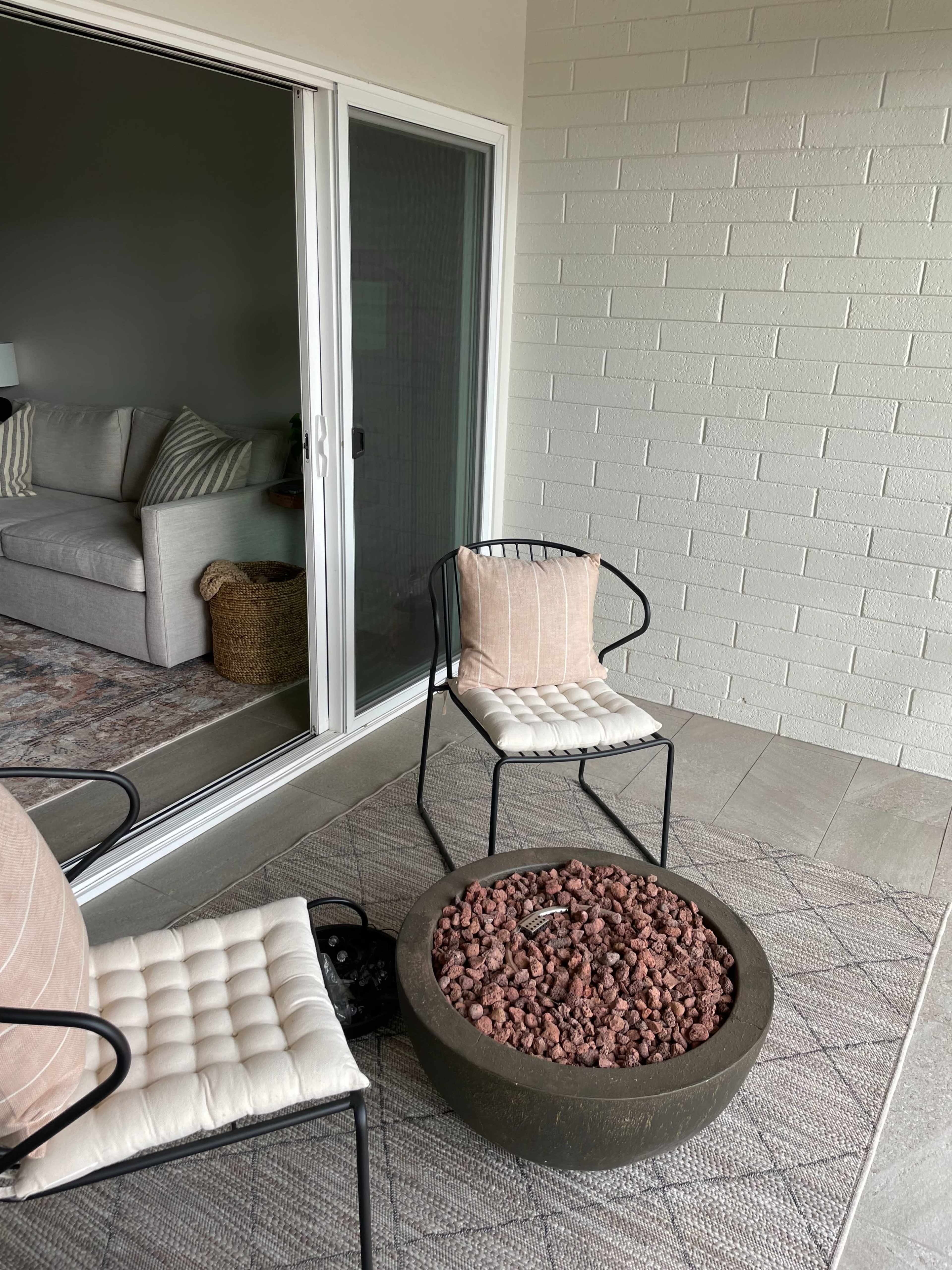 A small porch area with two black metal chairs facing a round concrete fire pit filled with decorative stones, next to a sliding glass door leading to an interior room.