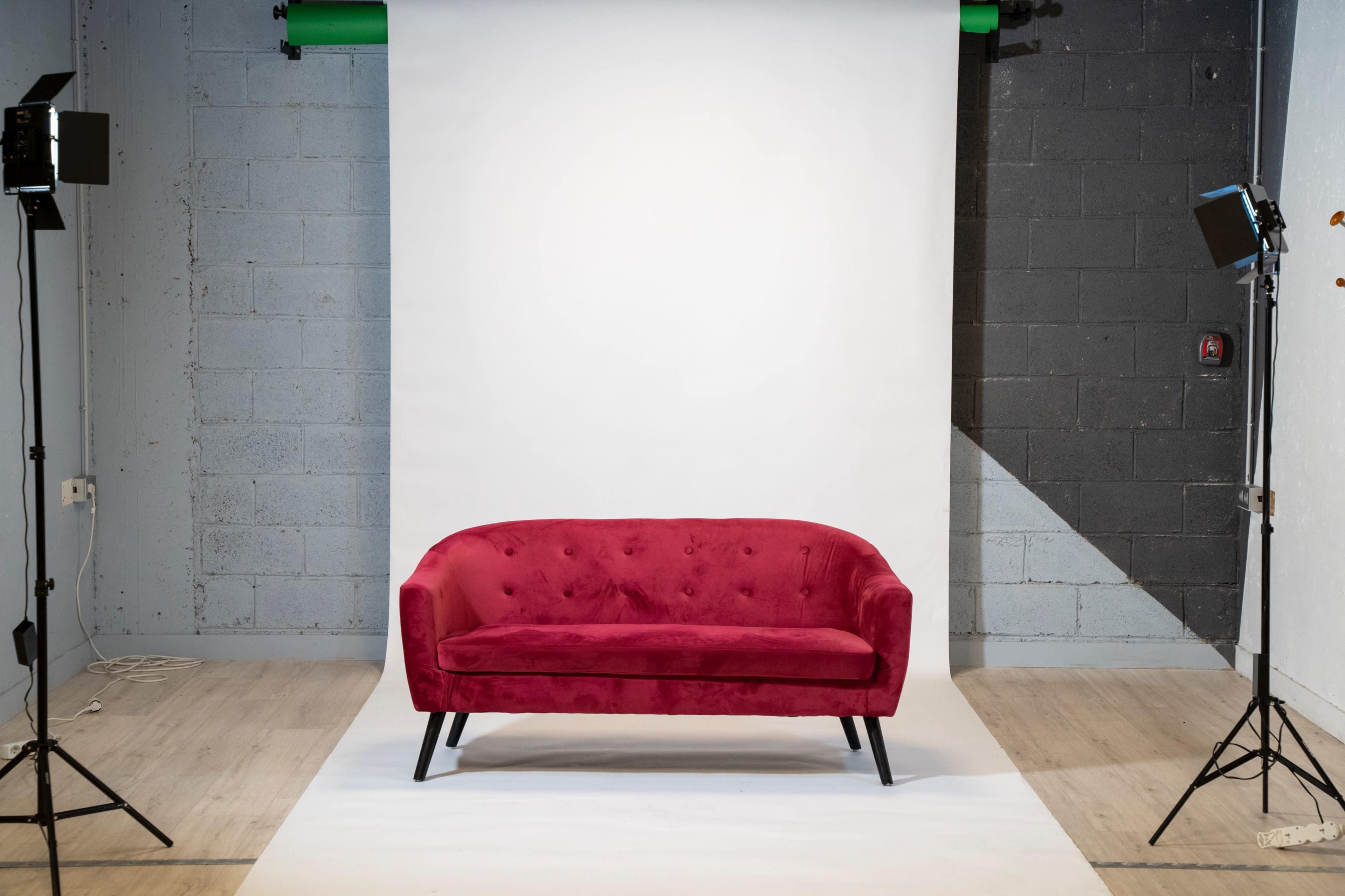 A red velvet sofa is centered in a photography studio with a white backdrop and two lighting setups on either side.