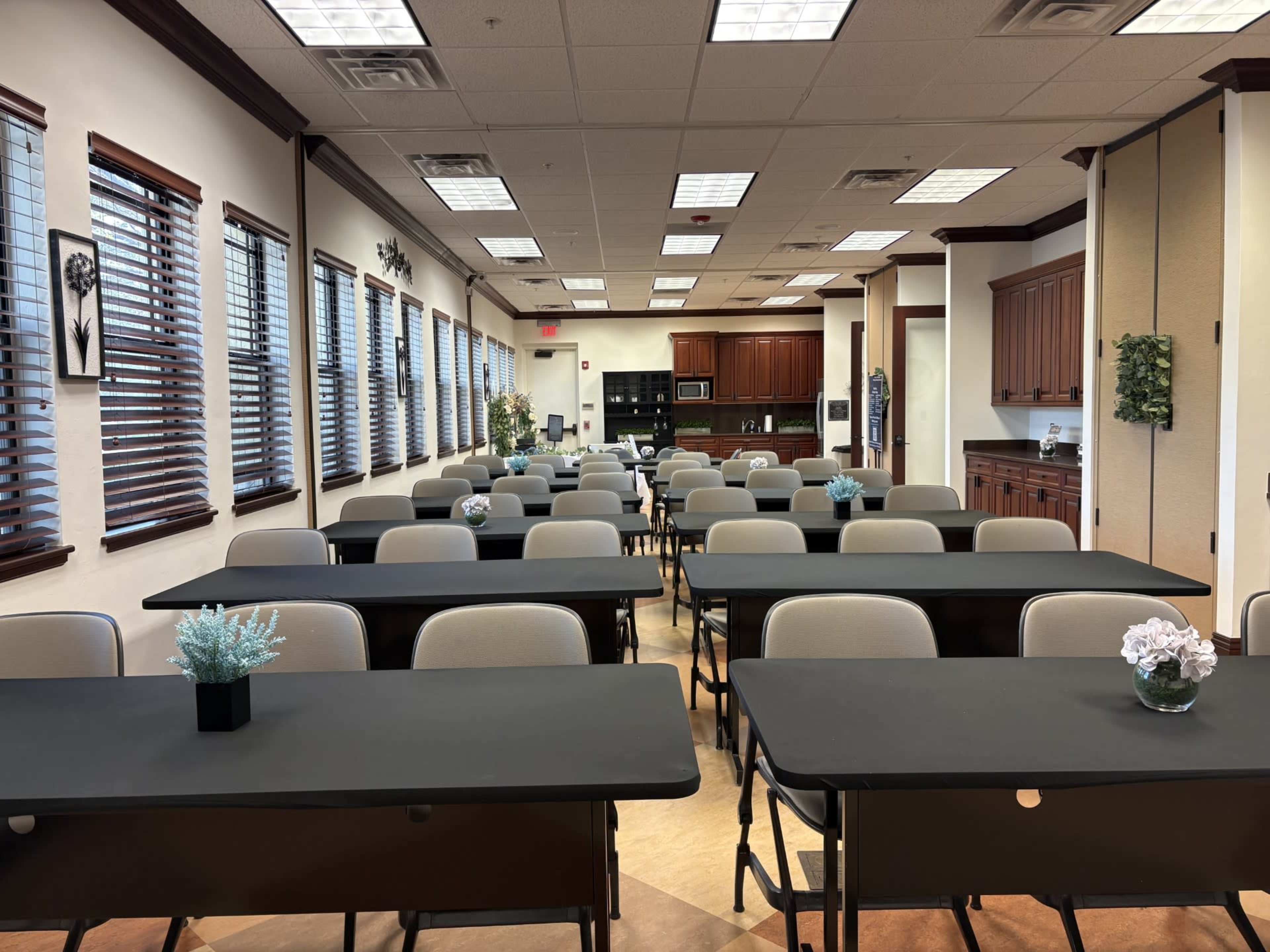 The image shows a spacious classroom setup with rows of tables and chairs, featuring a kitchenette area in the background and decorative plants on the tables.