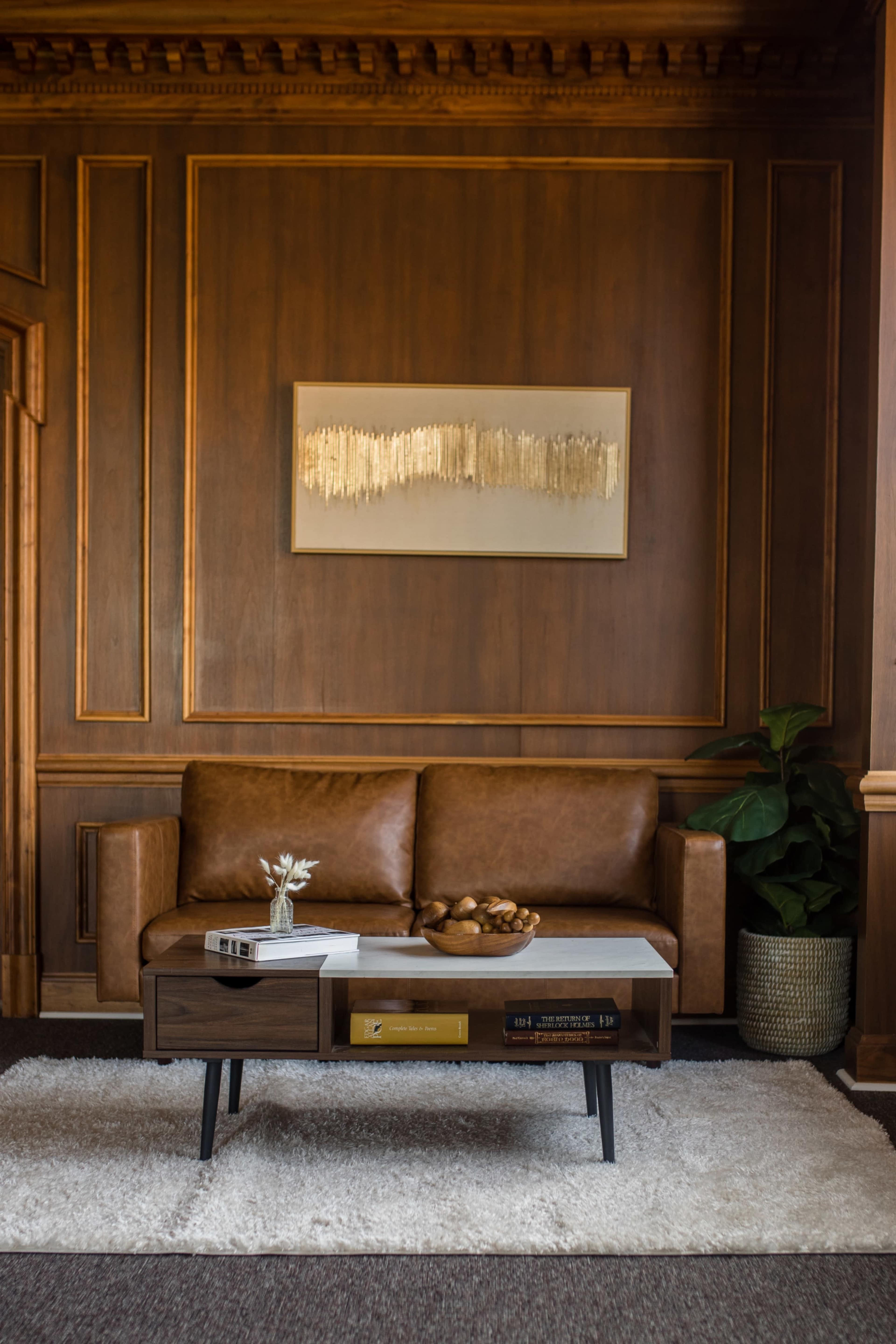A brown leather sofa is positioned against a wood-paneled wall, with a coffee table in front of it and a potted plant nearby.